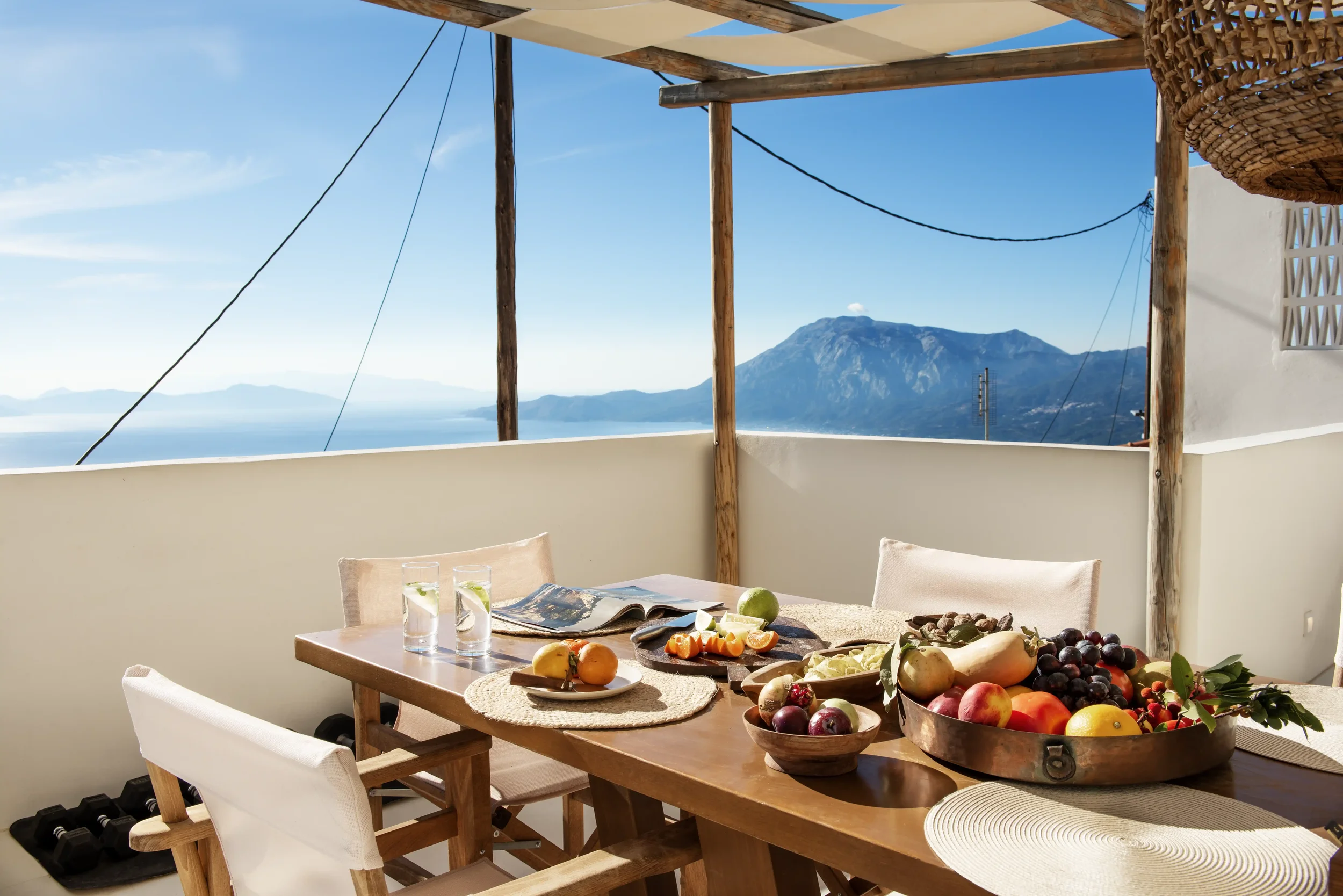 Outdoor dining area with a wooden table set with fresh fruit, glasses of water, and an open magazine under a pergola, overlooking a mountain and the sea.