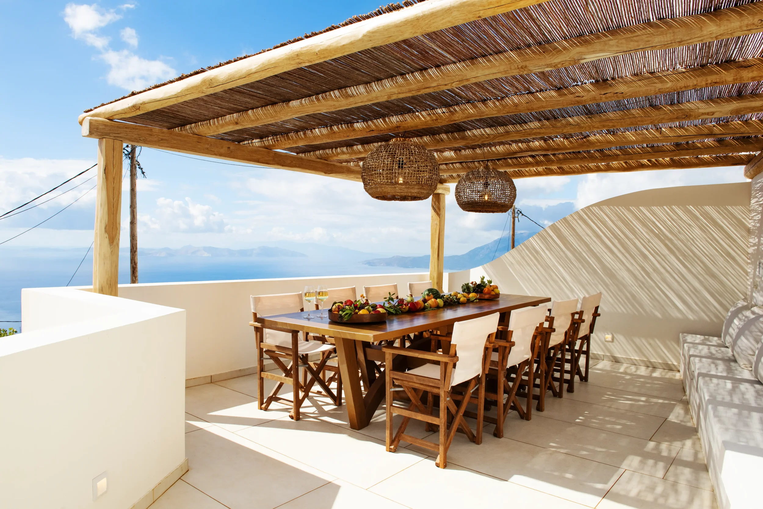 Outdoor terrace with wooden dining table, chairs, and fruit platter, overlooking the ocean with blue sky and clouds