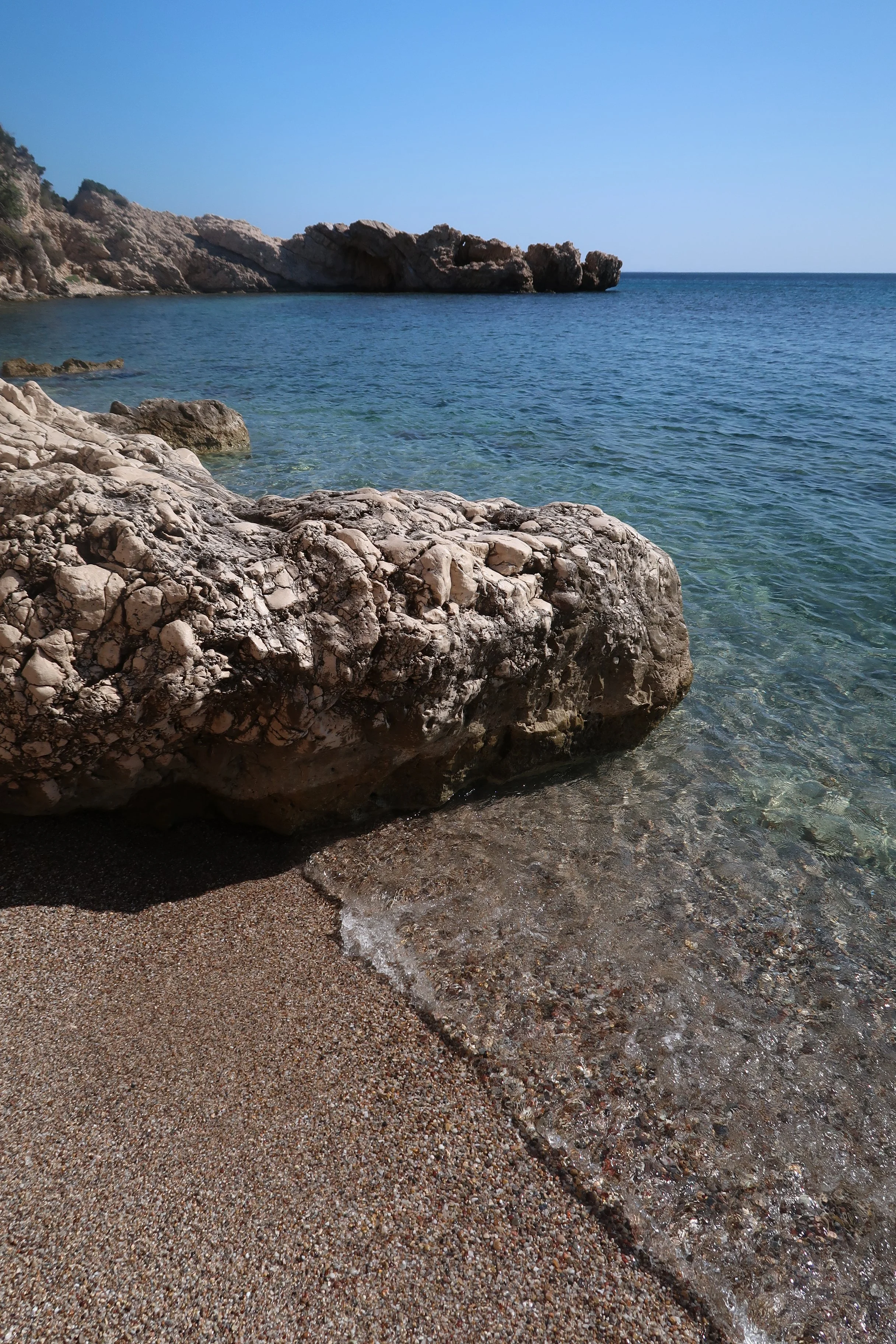 A rocky beach with clear water, a large rock in the foreground, and a cliff in the distance under a blue sky.