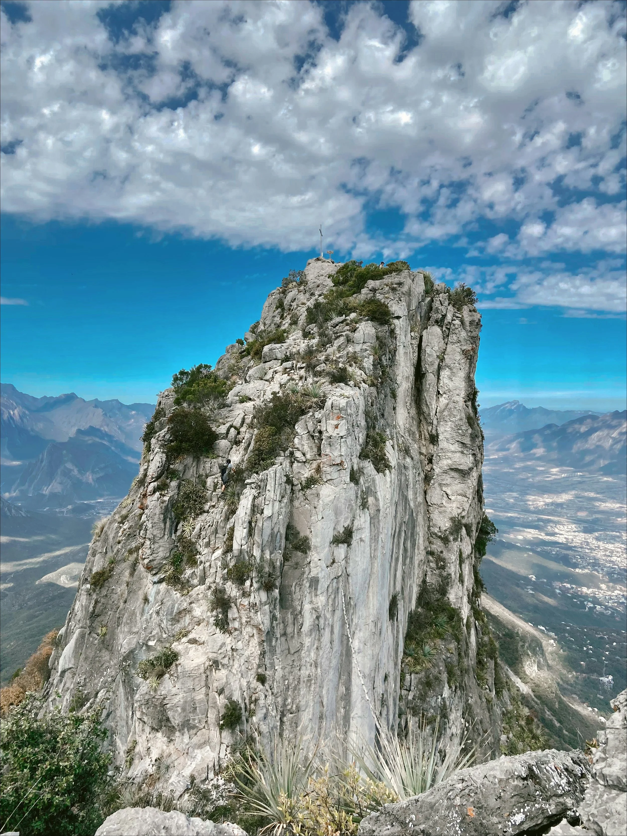 A tall rocky mountain peak with sparse vegetation and a flag at the summit, with a valley and mountain range in the background under a partly cloudy sky, in Samos