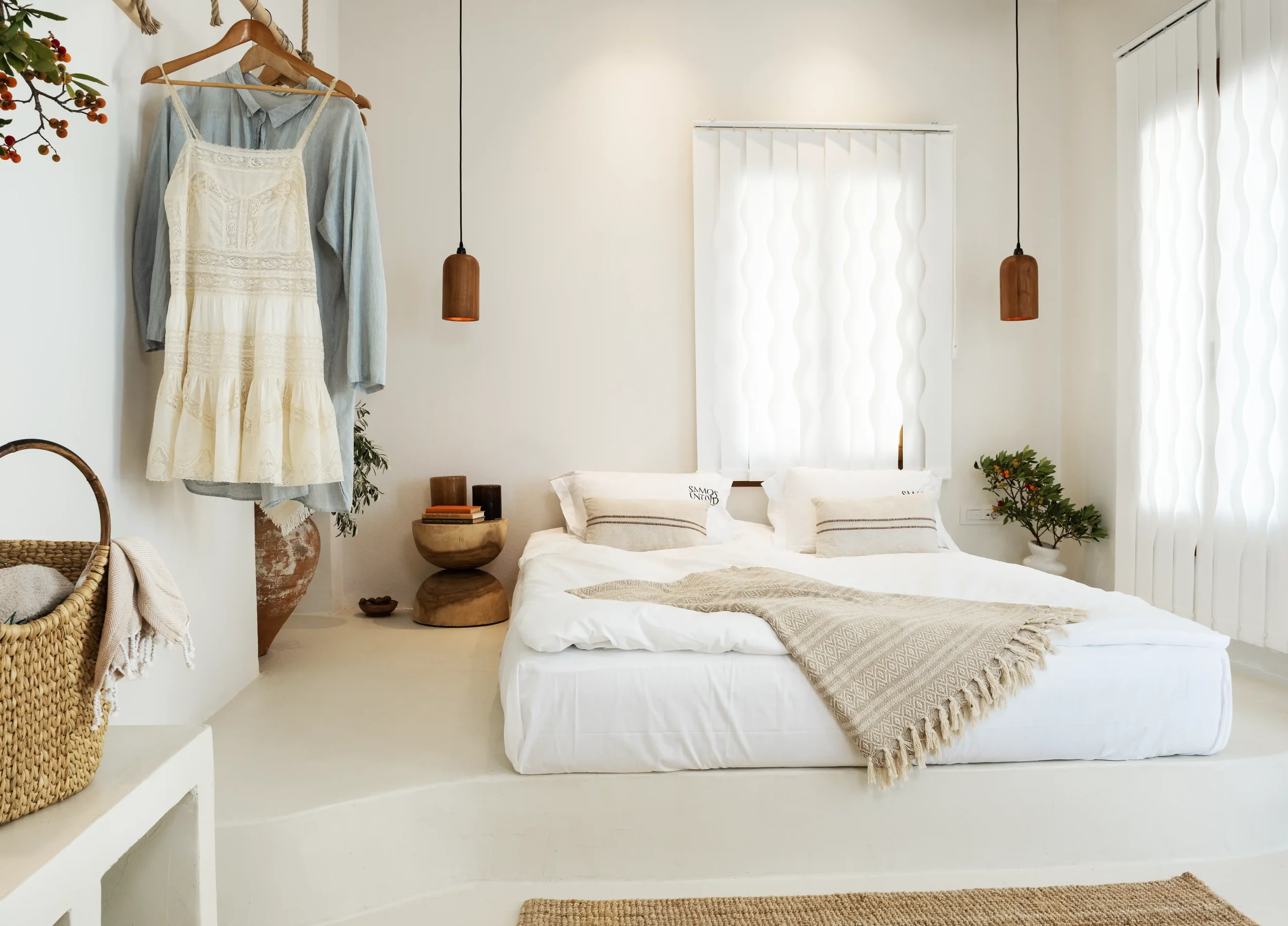 A minimalist white bedroom with a platform bed, hanging wooden pendant lights, and natural decor including plants, wicker basket, and a small nightstand with books. Light flows through vertical blinds on the windows.