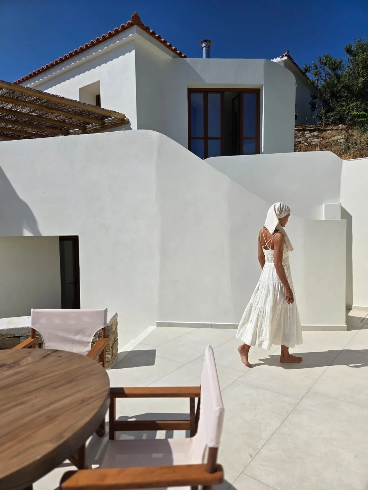 A woman in a white dress and head covering walking barefoot on a sunlit patio in front of white modern buildings with curved walls, wooden table, and chairs.