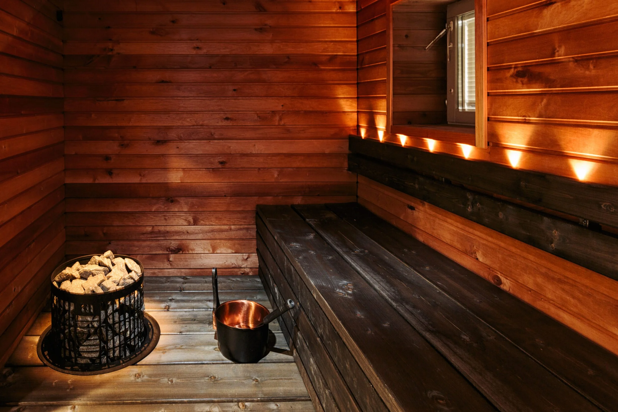 Huohetuinen sauna sisällä, puupaneelit ja lauteet, kiuaskiviä ja sanko, valaistus.