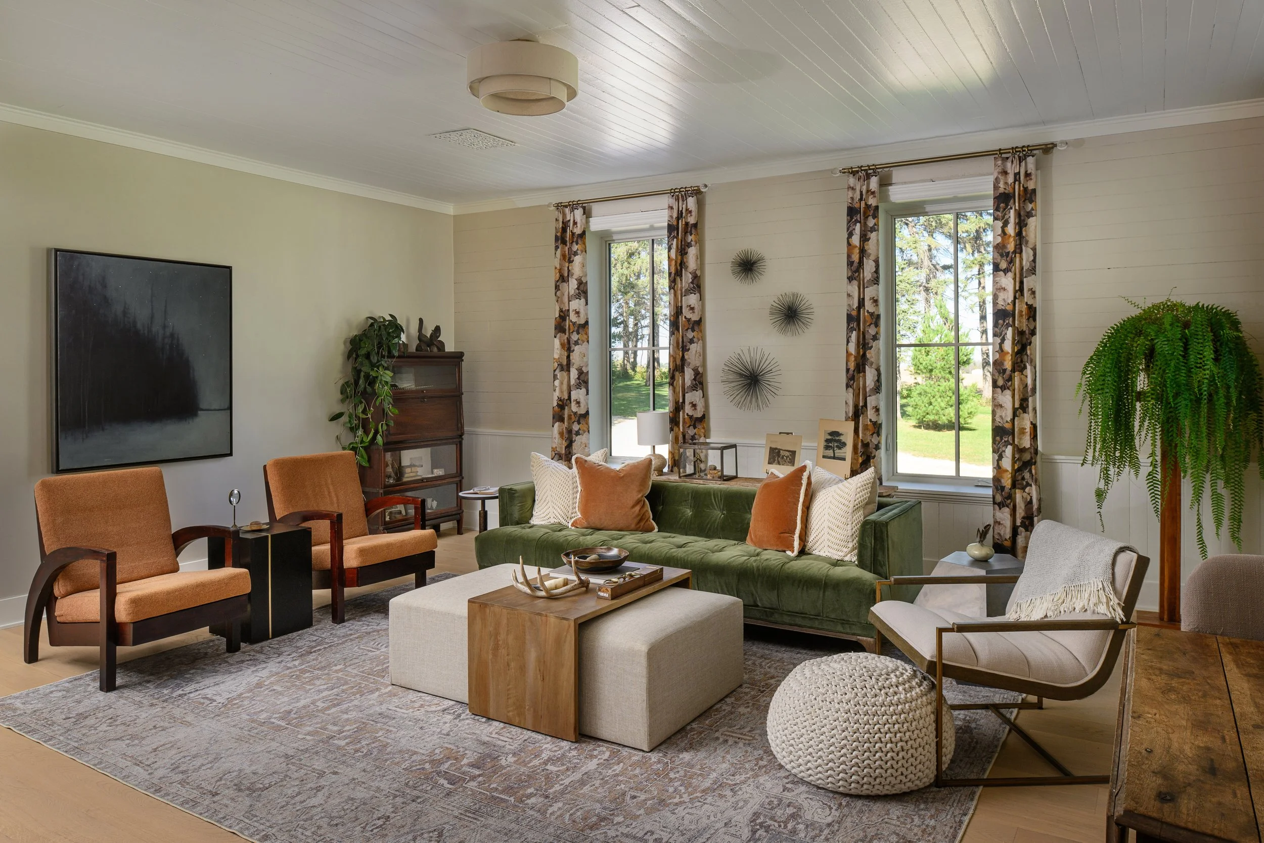 Living room with green velvet sofa, two orange armchairs, a white pouf, a wooden coffee table, and various decorative items, windows with floral curtains, and houseplants.