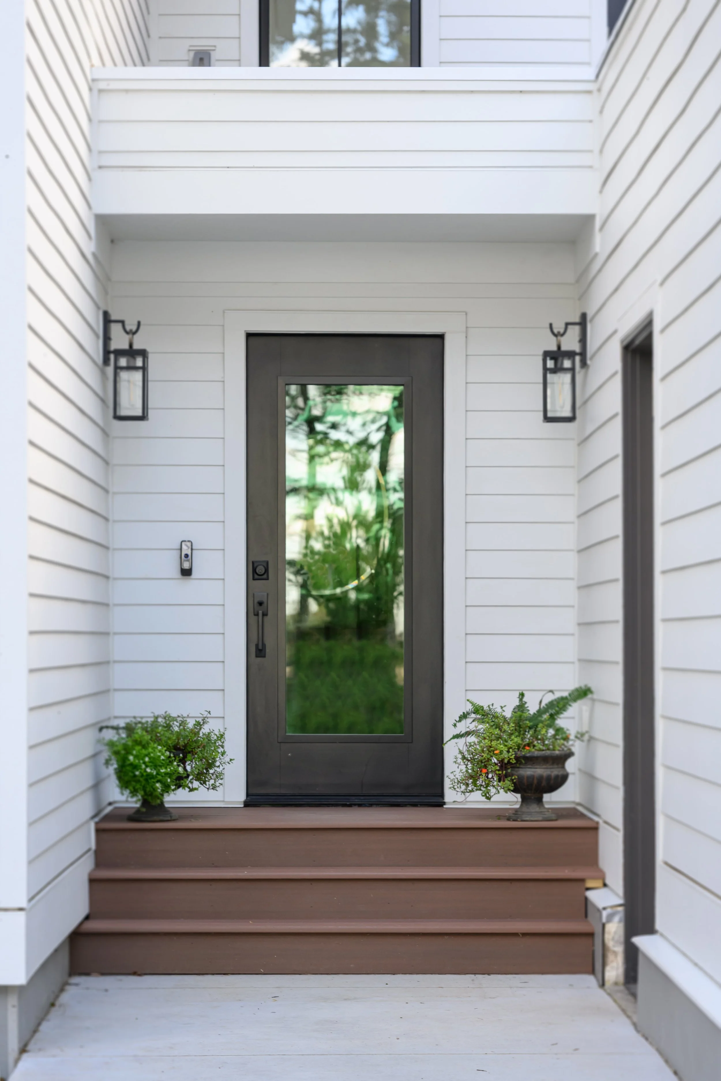 Modern front door with glass panel, flanked by two wall-mounted lanterns, with potted plants on steps leading to the entrance, white siding on house exterior.