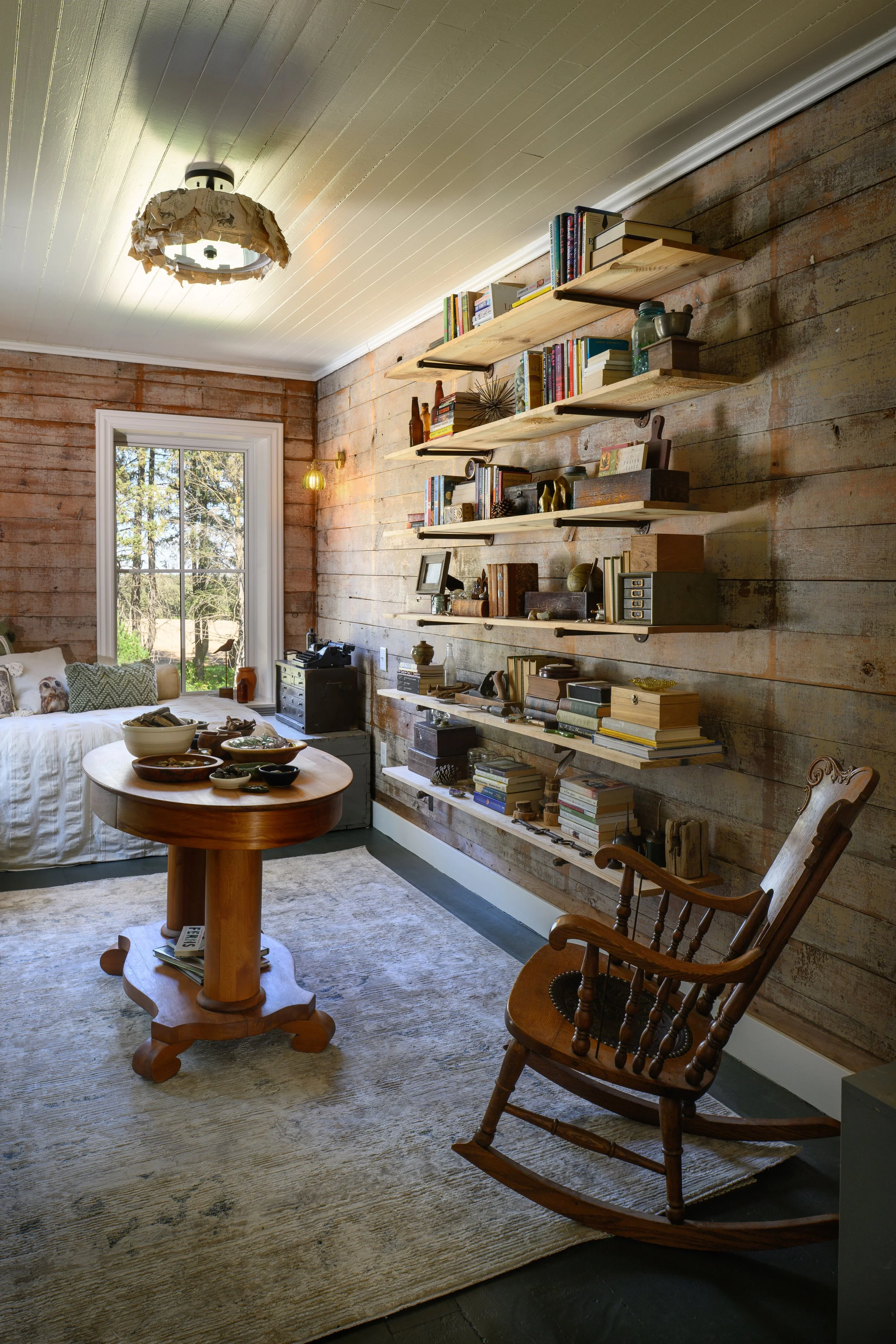 A cozy living room with a rustic wooden wall, a window with a view of trees, a round wooden table, a rocking chair, and floating shelves filled with books and decorative items.