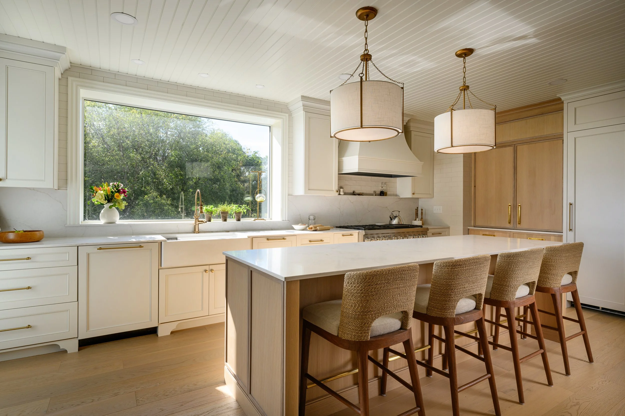 Modern kitchen with white cabinetry, a large window, a central island with beige chairs, pendant lights, and a view of greenery outside.