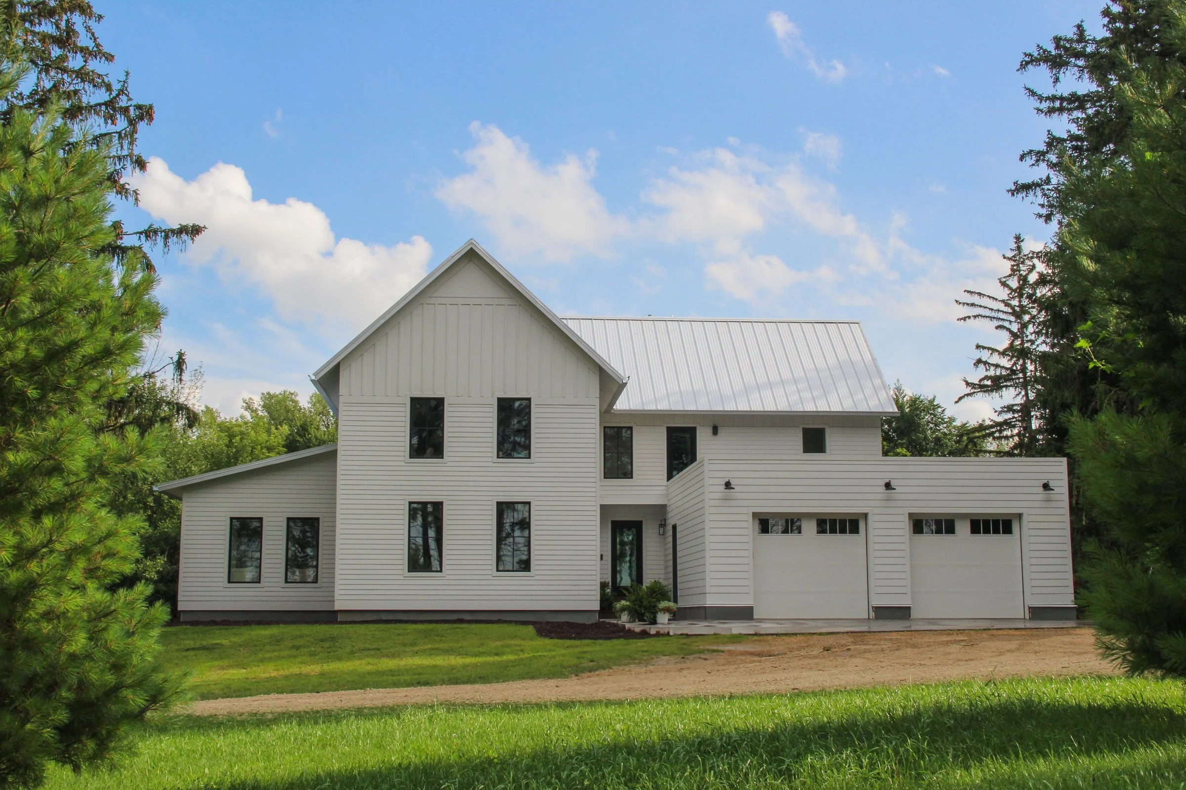 White modern house with metal roof, multiple windows, two-car garage, surrounded by grass and trees, under a partly cloudy sky.