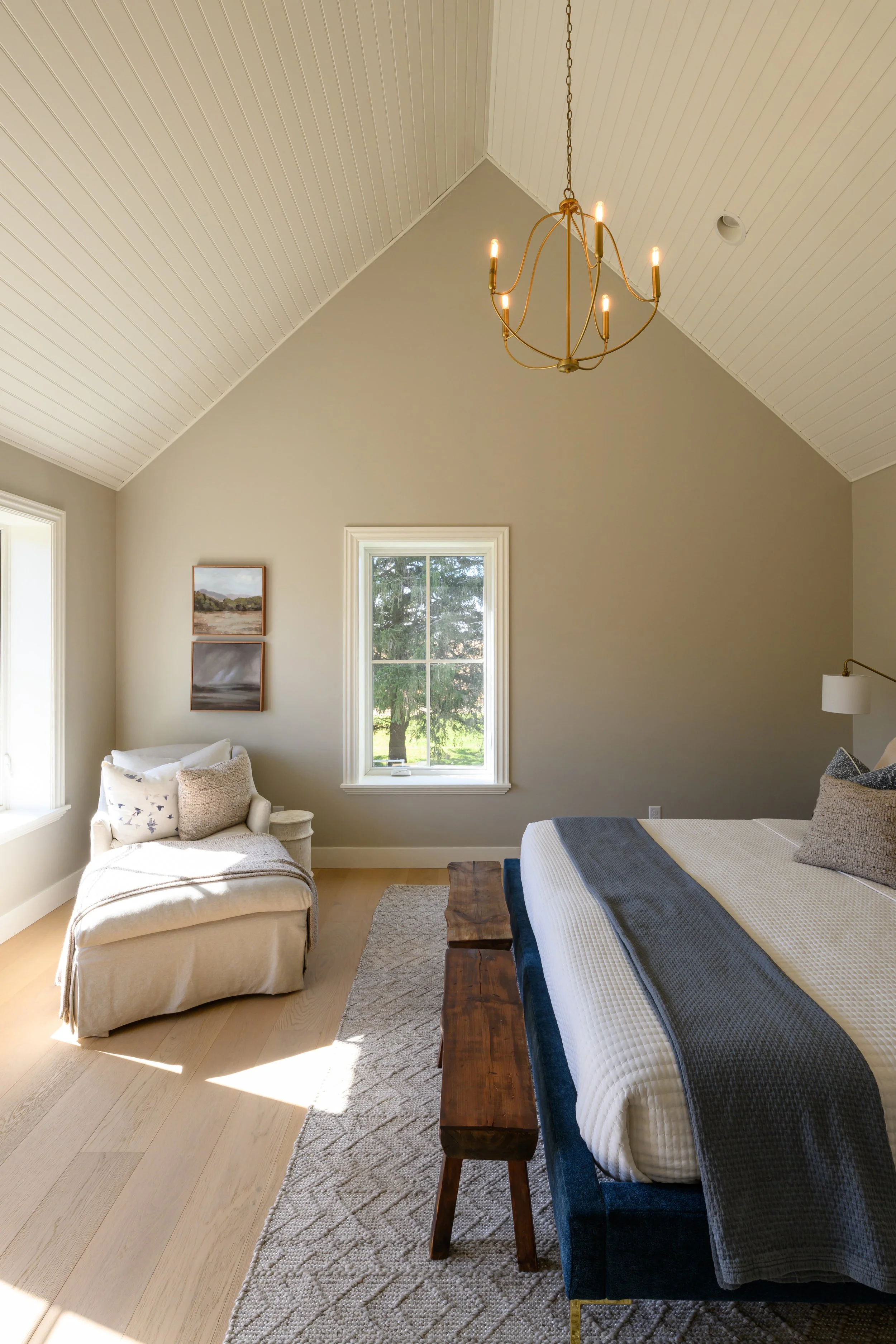 A bedroom with a vaulted ceiling, a chandelier, a window showing greenery outside, a bed with pillows and a blanket, a wooden bench at the foot of the bed, a beige armchair with pillows, and framed artwork on the wall.