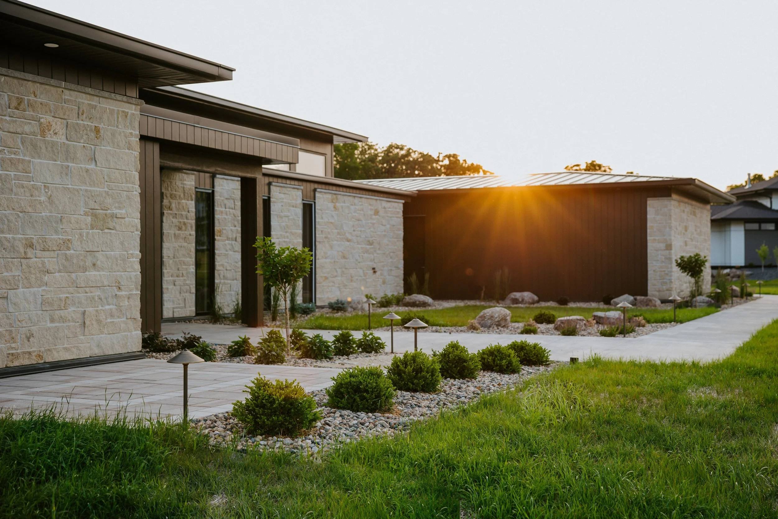 Modern house with Edwards Cottonwood Dimensional stone and Zola Windows, landscaped front yard with small bushes, rocks, trees, pathway, and outdoor lights at sunset.
