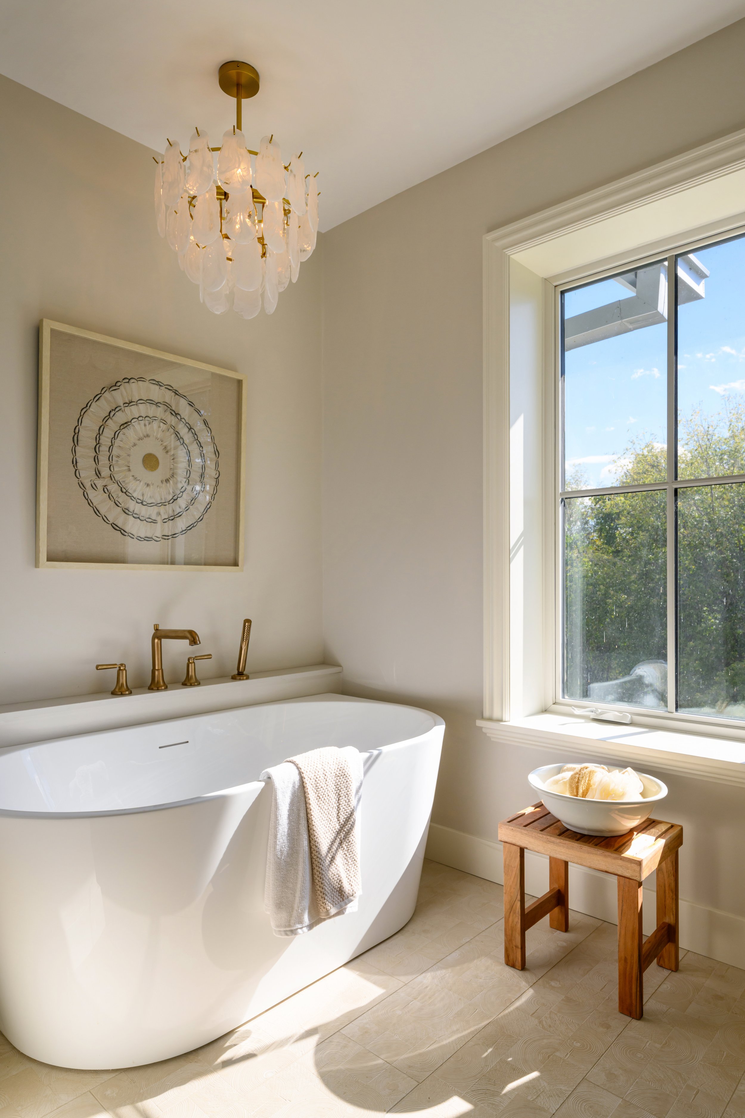 A bathroom with a freestanding bathtub, a large window showing a view of trees, a wooden stool with a white bowl, a framed artwork on the wall, and a decorative chandelier hanging from the ceiling.