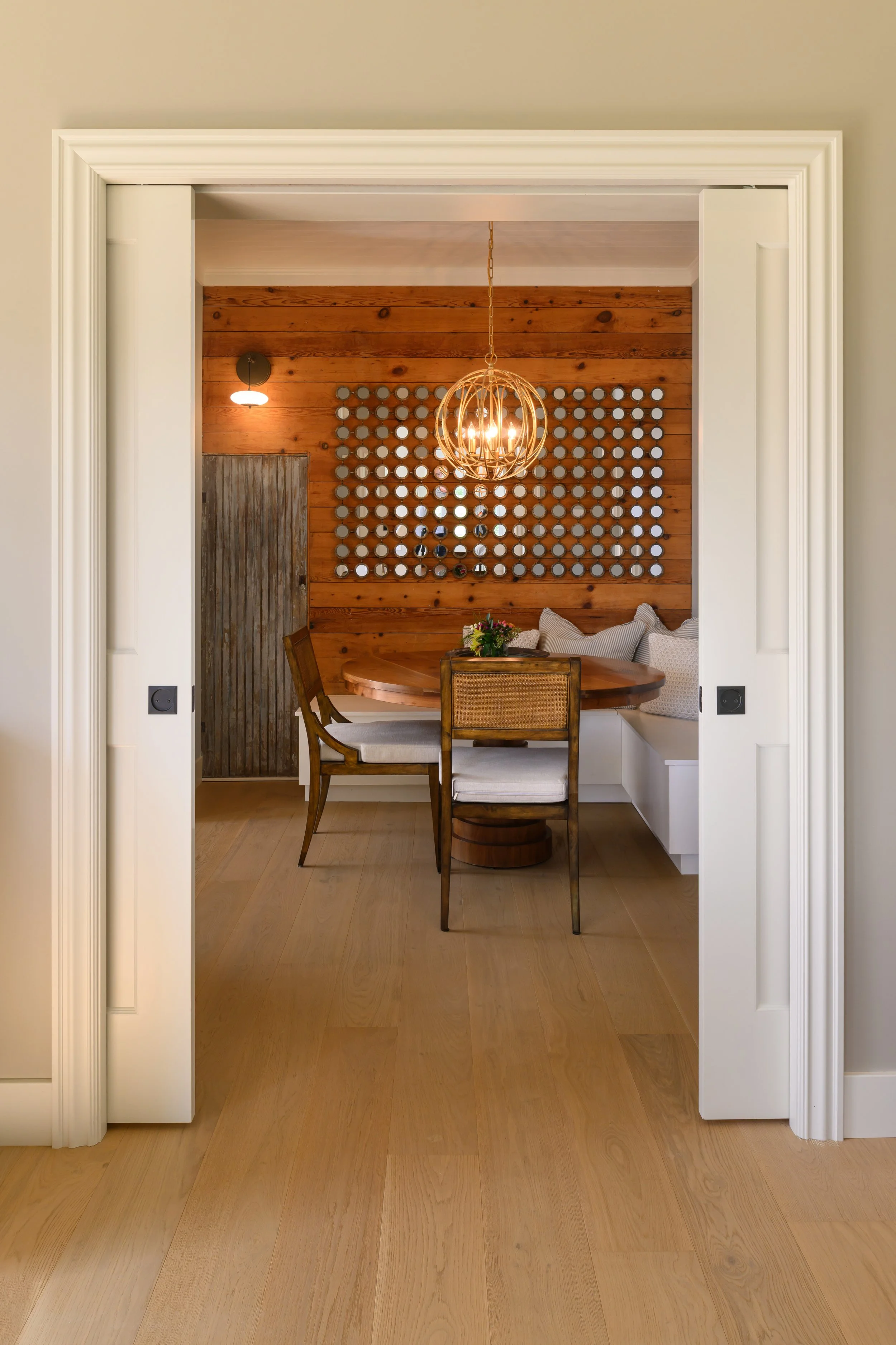 View of a cozy dining nook with a round wooden table, chairs, and a built-in bench with cushions, set against a wooden wall with decorative round mirrors and a distinctive hanging light fixture.