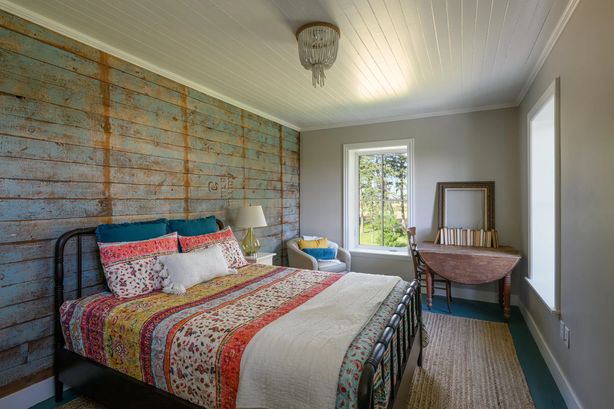 Bedroom with a bed with colorful bedding and pillows, a white armchair with a yellow pillow, a wooden desk with stacked books, a window showing greenery outside, and a rustic wooden accent wall.