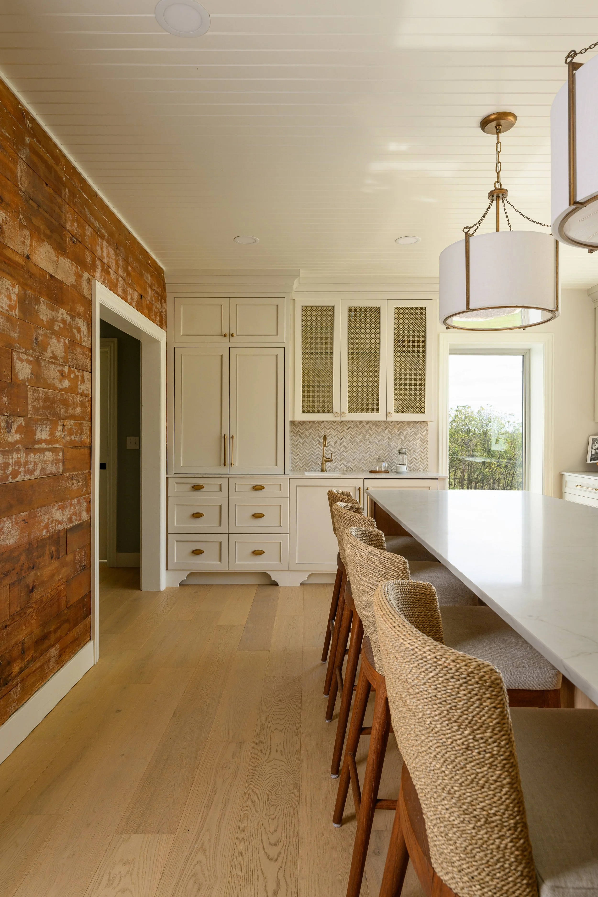 A kitchen with a white island counter, woven bar chairs, white cabinetry, a wood accent wall, and a large window showing green trees outside.