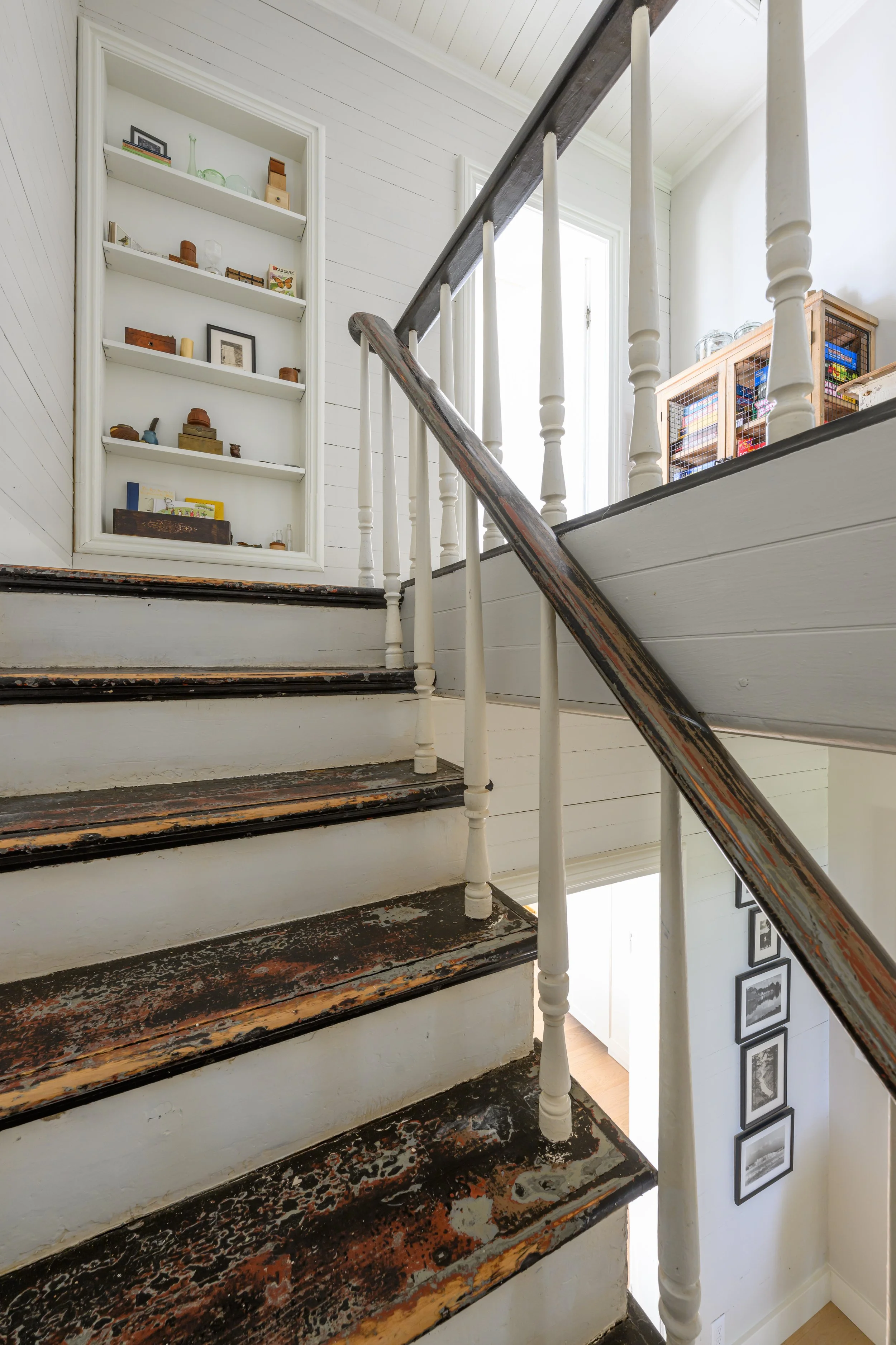 Interior view of a staircase with worn, chipping black and white paint, leading upward next to a white wall with picture frames. A window and a built-in bookshelf filled with decorative items and books are visible in the background.