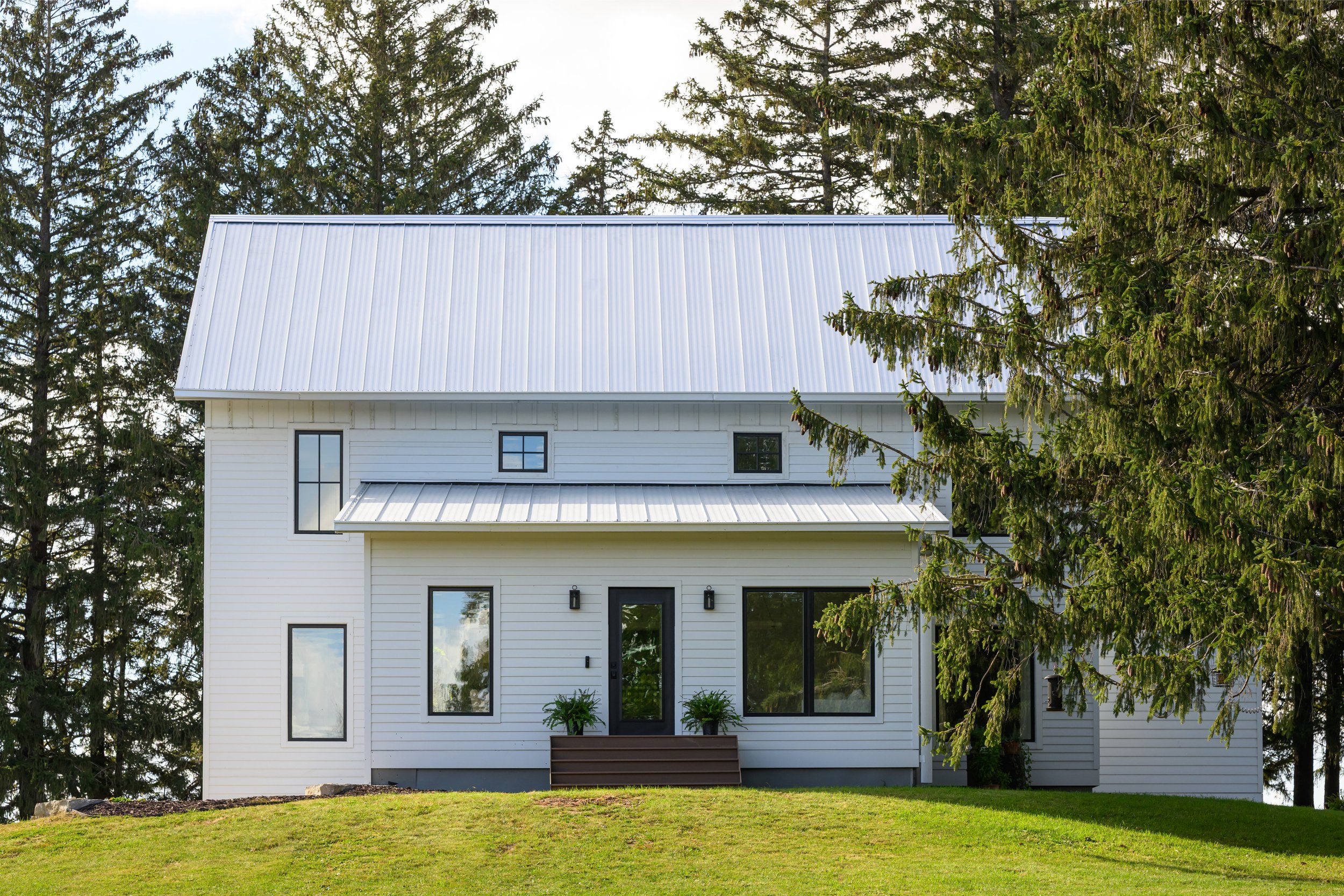 A modern white house with black window frames and a metal roof, surrounded by green grass and tall trees.