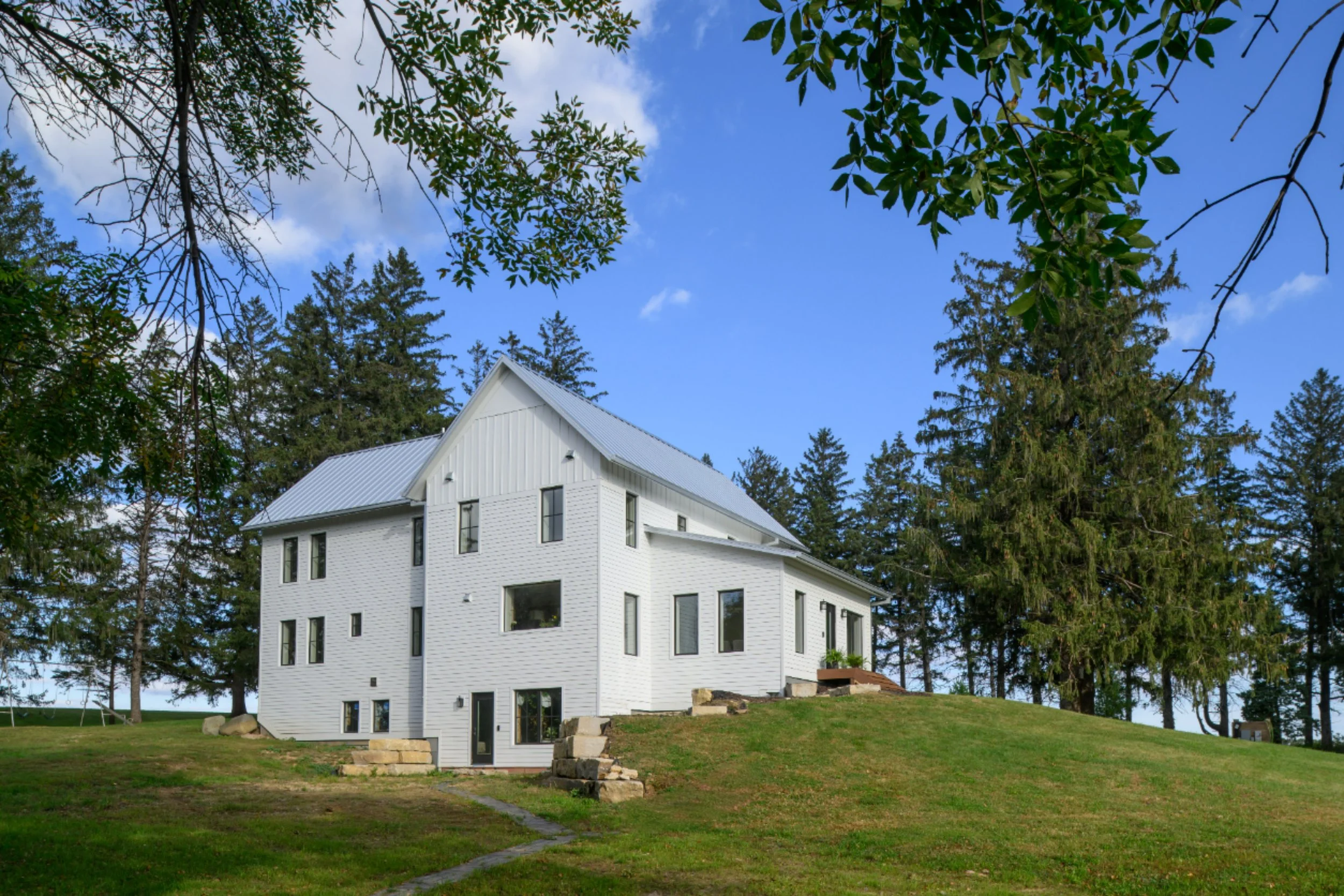 A white multi-story house on a grassy hill surrounded by tall trees with a blue sky overhead.
