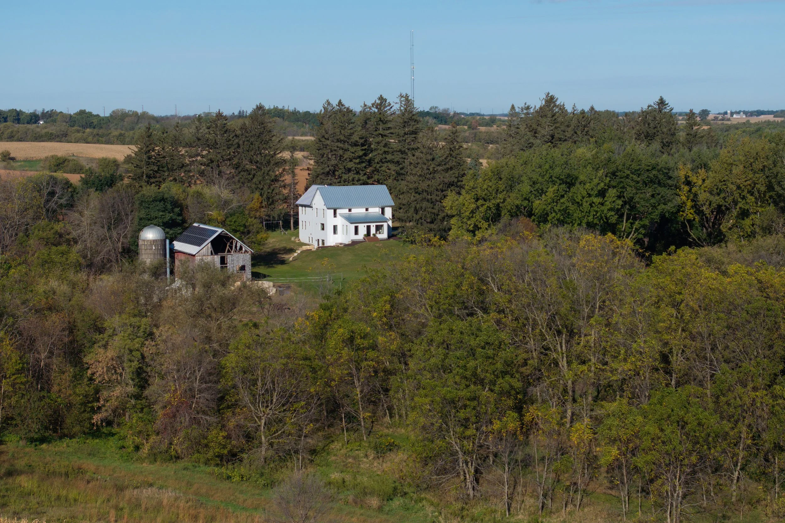 A rural landscape with a white house, a barn with solar panels, and a silo surrounded by trees and fields under a clear blue sky.