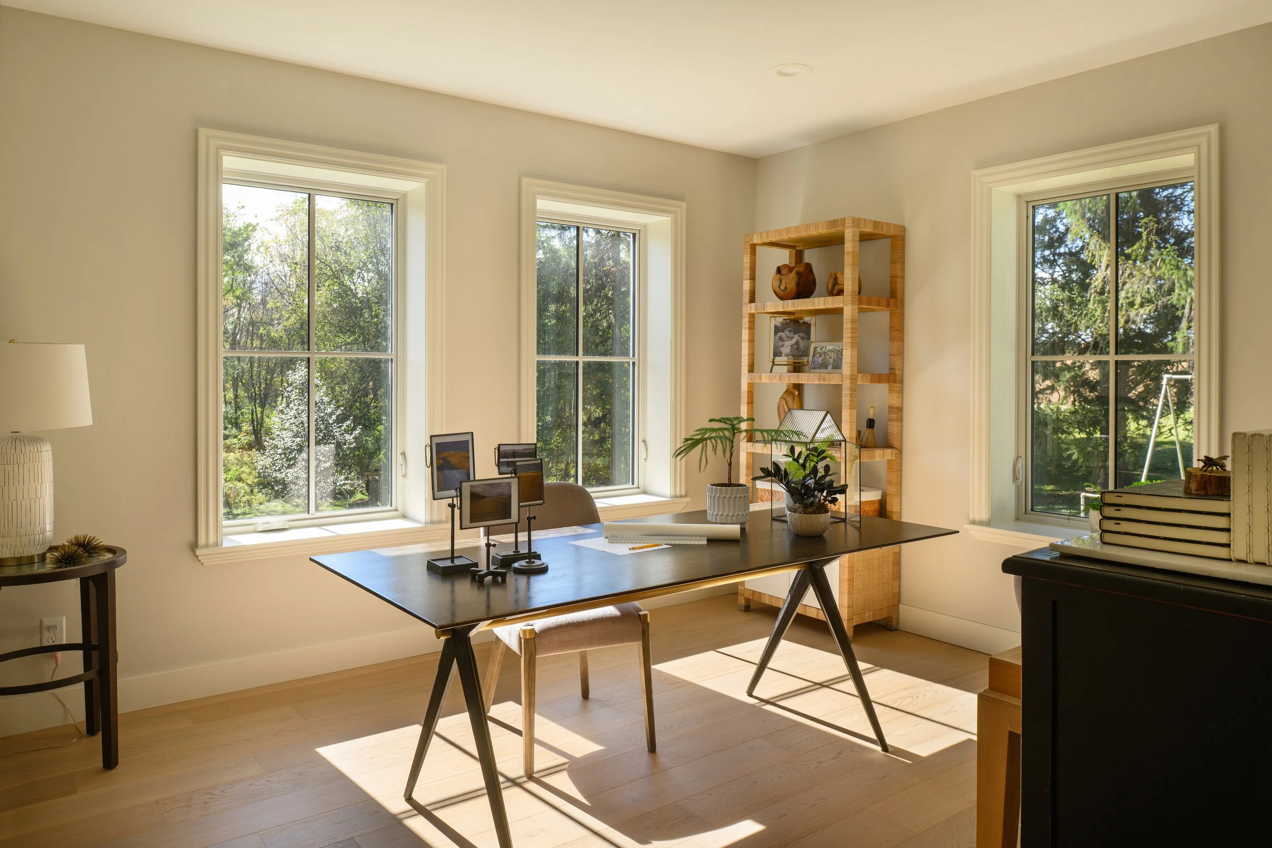 Bright home office with three large windows, a dark desk with decorative plants and framed artwork, a wooden bookshelf with decor items, and a side table with books and a lamp.