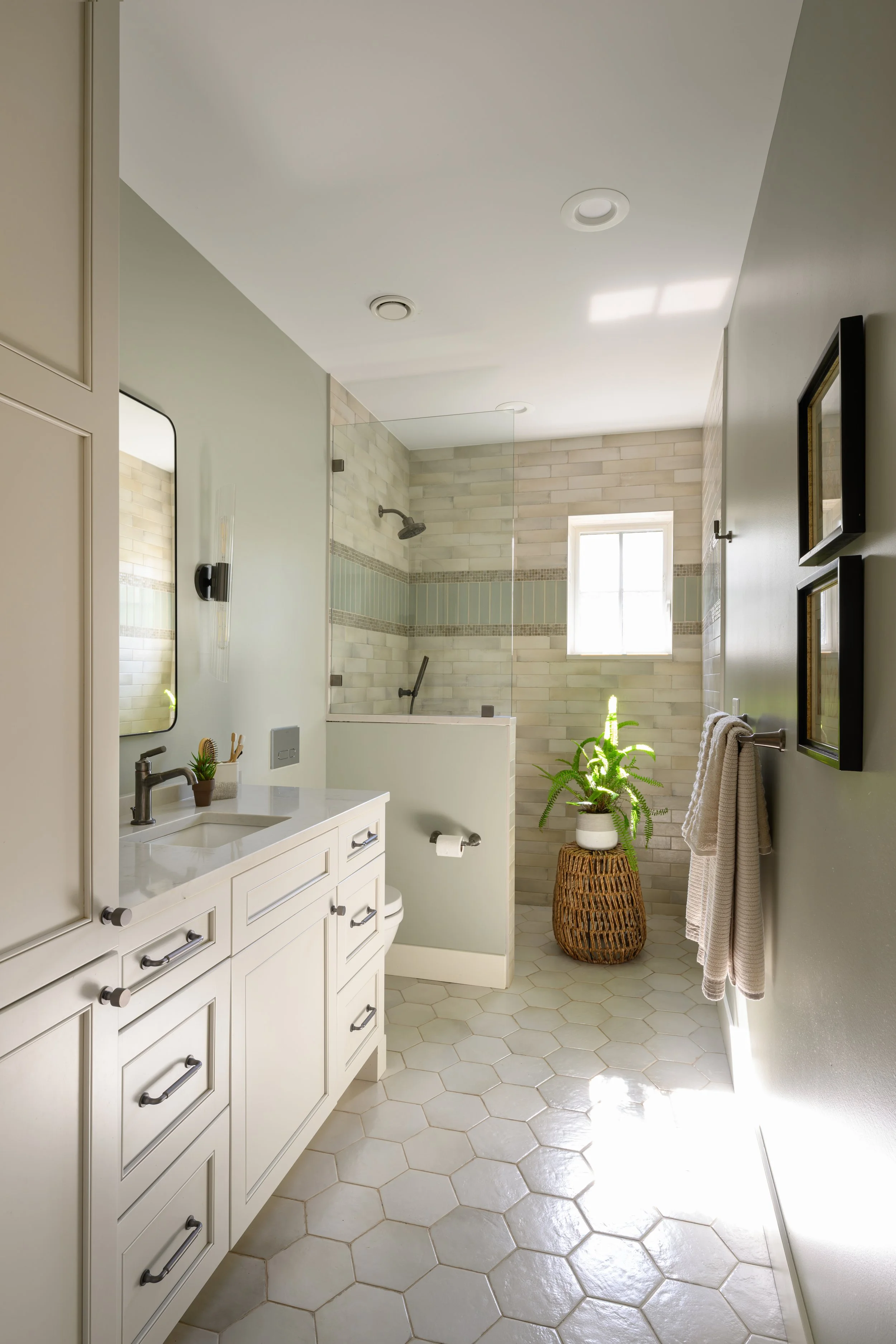 Bright bathroom with white vanity, round black handle drawer pulls, wall mirror, small potted cactus, and white sink. Shower area with glass partition, beige tile walls, small window, green potted plant on wicker stand, and beige hexagonal tiled floo