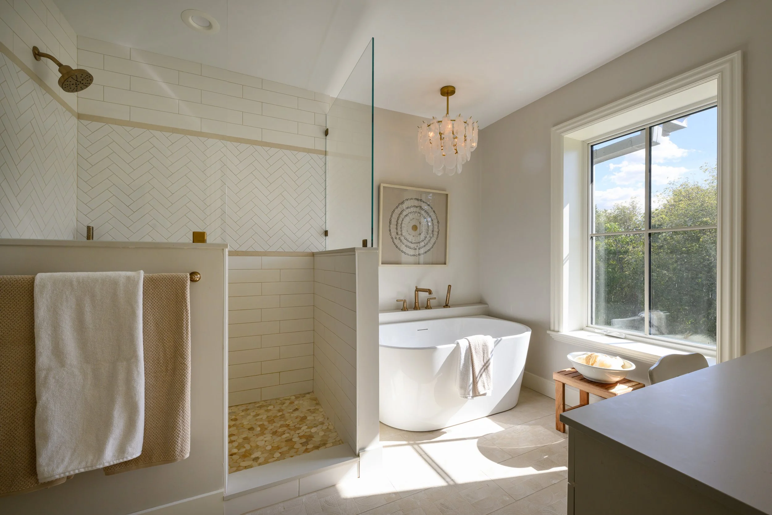 Bright bathroom with a large window showing trees outside, a white freestanding bathtub with gold fixtures, a walk-in shower with white herringbone and subway tile, a towel hanging on a rack, and minimal decor including a chandelier and wall art.