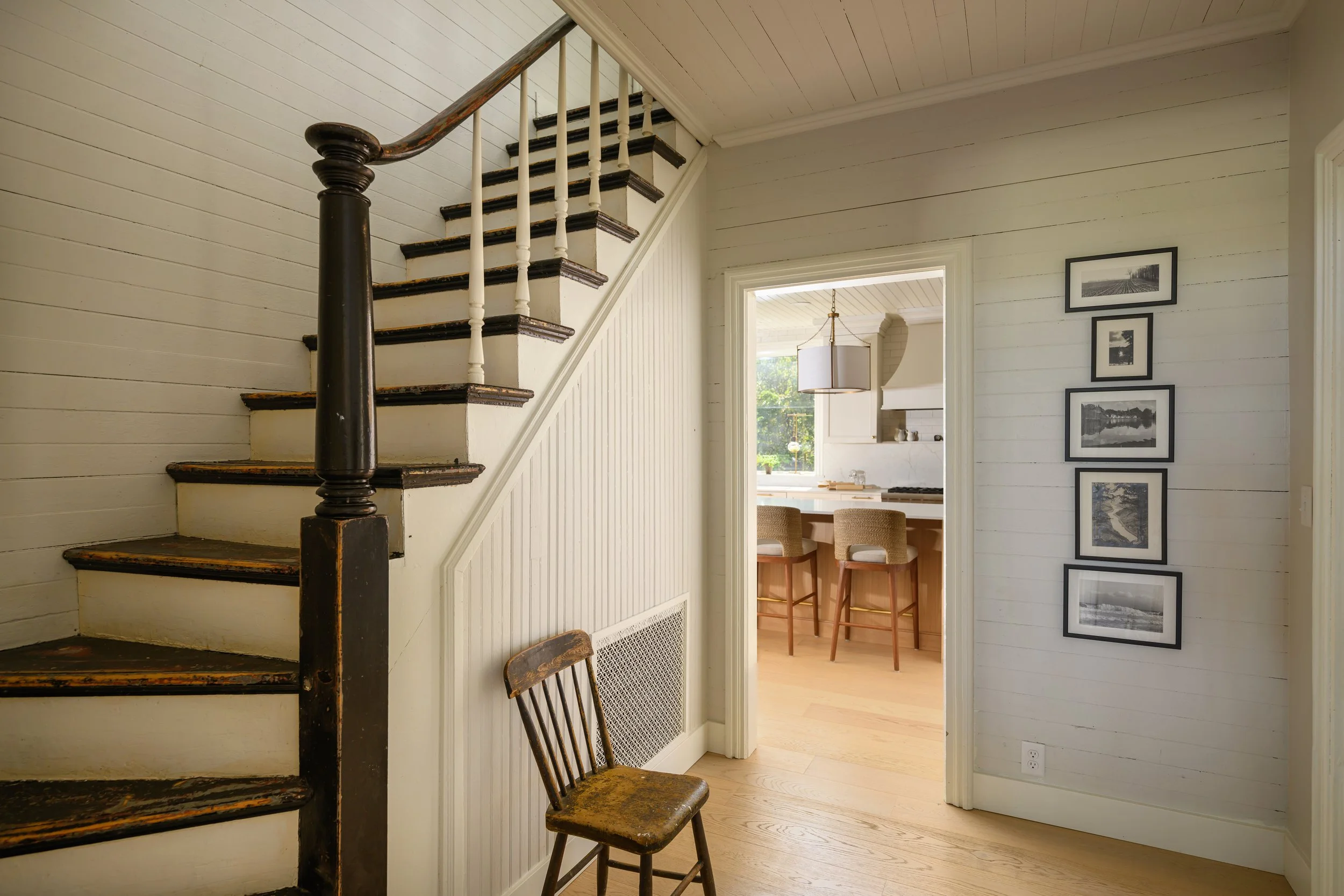 Interior view of a home showing a wooden staircase with black accents, a chair, a doorway leading to a kitchen with bar stools, and framed photos on the wall.