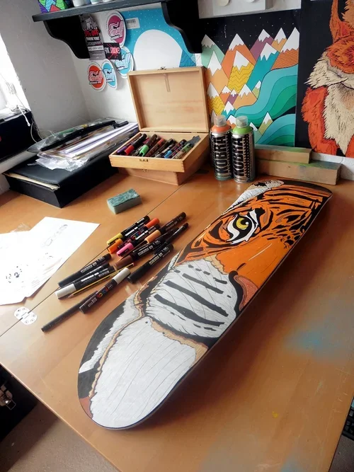 A skateboard deck with a tiger face design in progress, accompanied by markers, spray paints, and art supplies on a wooden table.