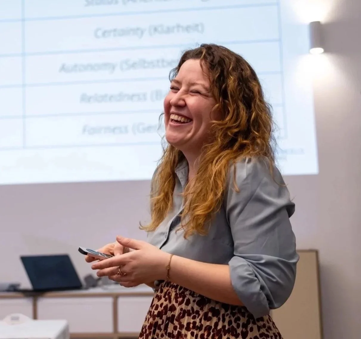 Frau in weißem T-Shirt und gemustertem Rock, lächelnd, mit verschränkten Armen vor weißem Hintergrund.