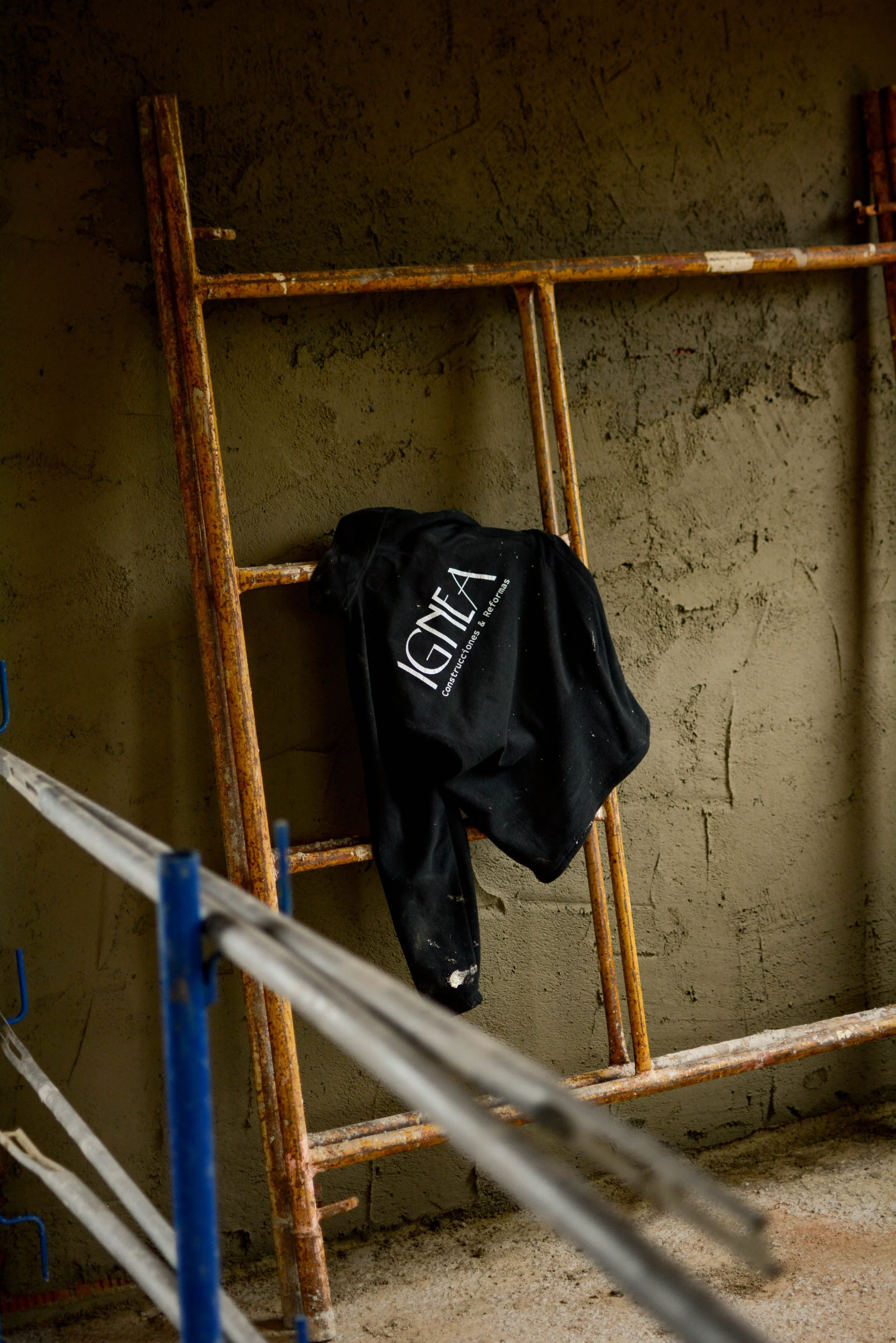 Escalera de andamio oxidado y camiseta negra colgada en ella, en un muro de concreto en construcción. Logotipo diseñado por GustoStudio