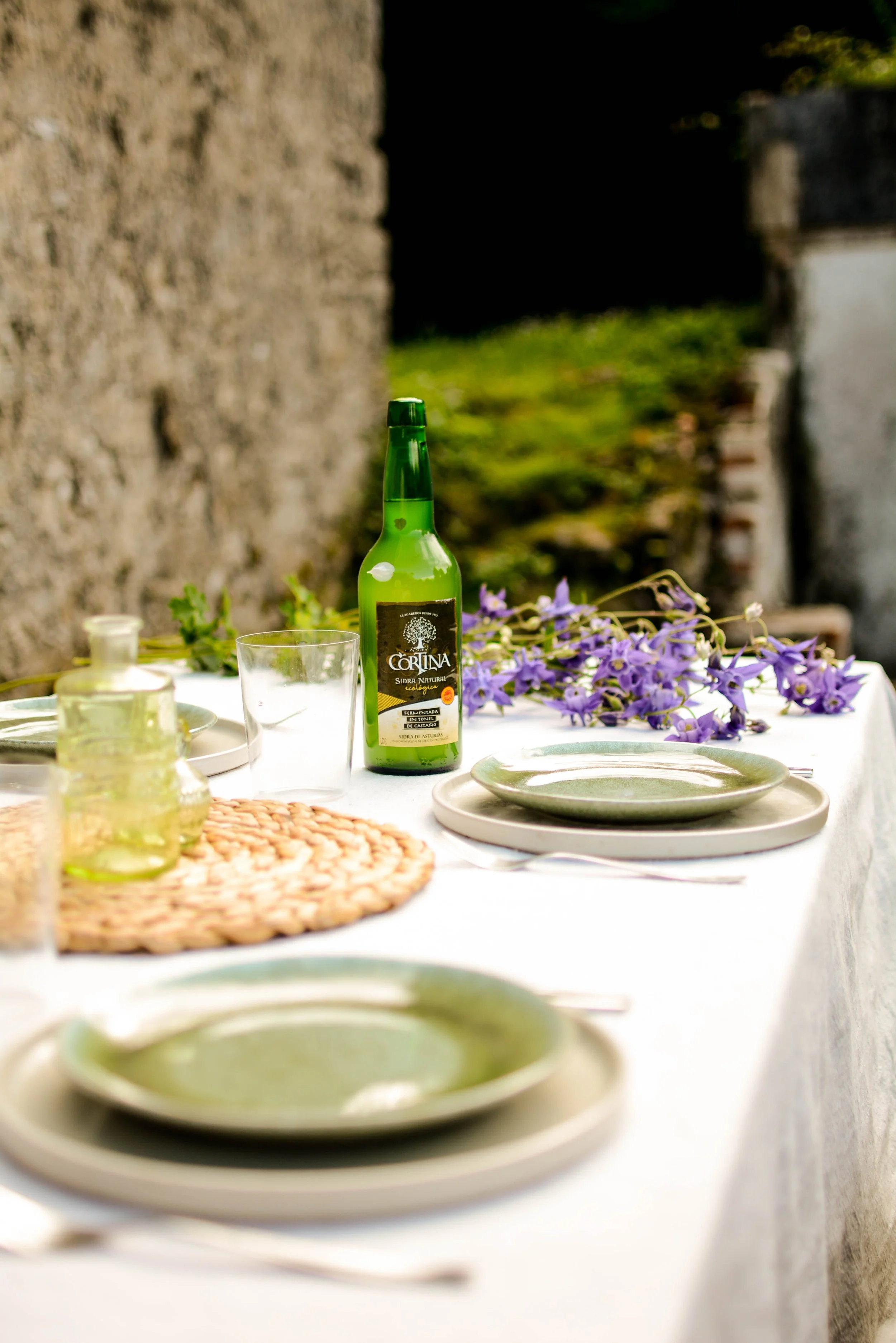 Mesa al aire libre con platos verdes, una botella de sidra y flores violetas para la empresa de Cortina S.L. Dirección creativa y foto por GustoStudio. 