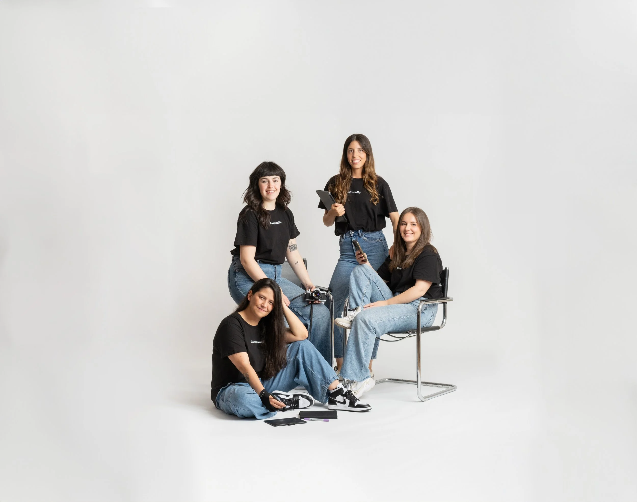 Equipo de GustoStudio al completo, somos cuatro jóvenes mujeres. Una está sentada en el suelo con una cámara y accesorios, otra sentada en una silla con un teléfono móvil, una más de pie con un cuaderno y teléfono, y la cuarta, sosteniendo una tablet