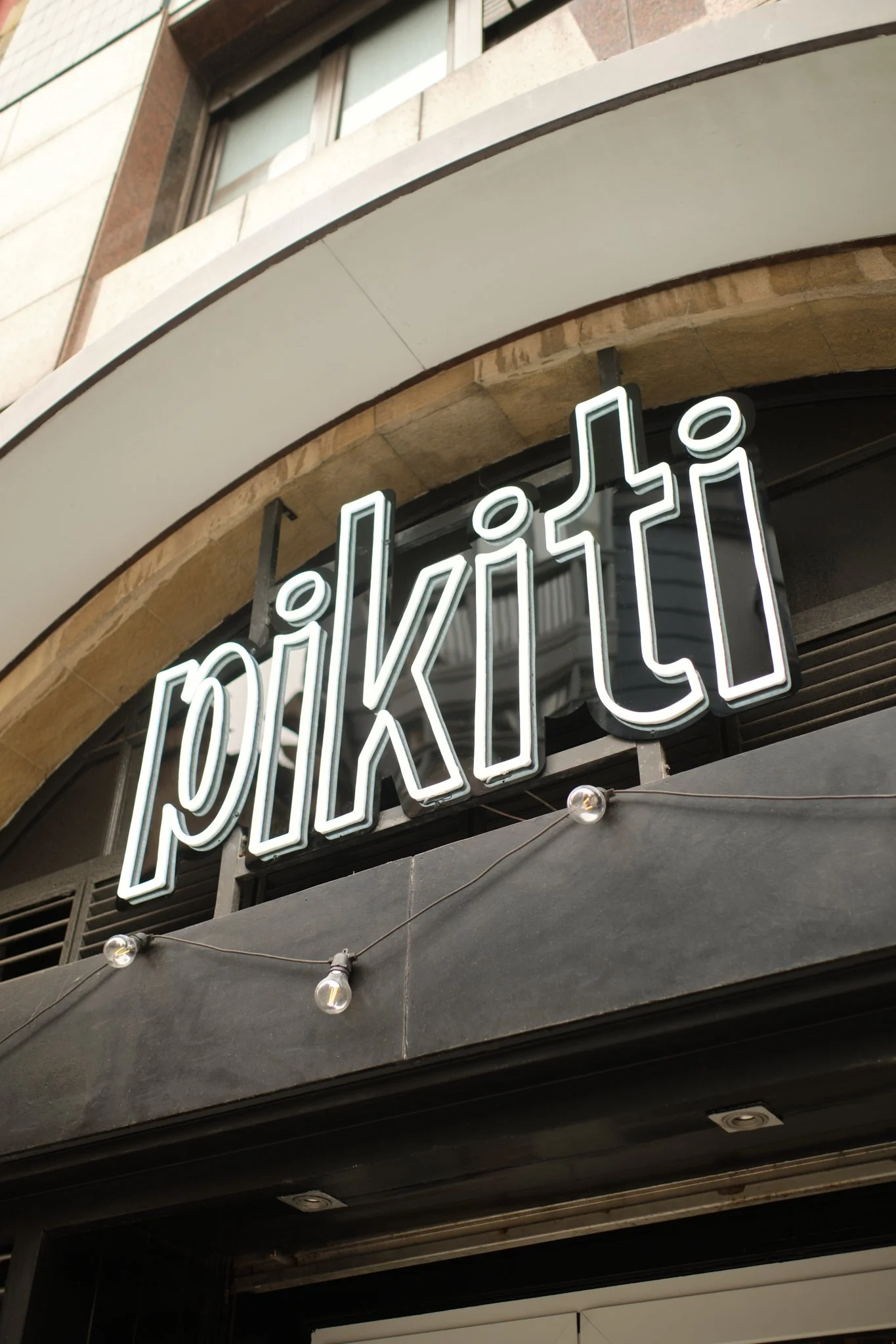 Logotipo que diseñamos para unos clientes en Gijón, Asturias. Cartel luminoso que dice 'pilikiti' en letras blancas con borde negro, en la fachada de un edificio.