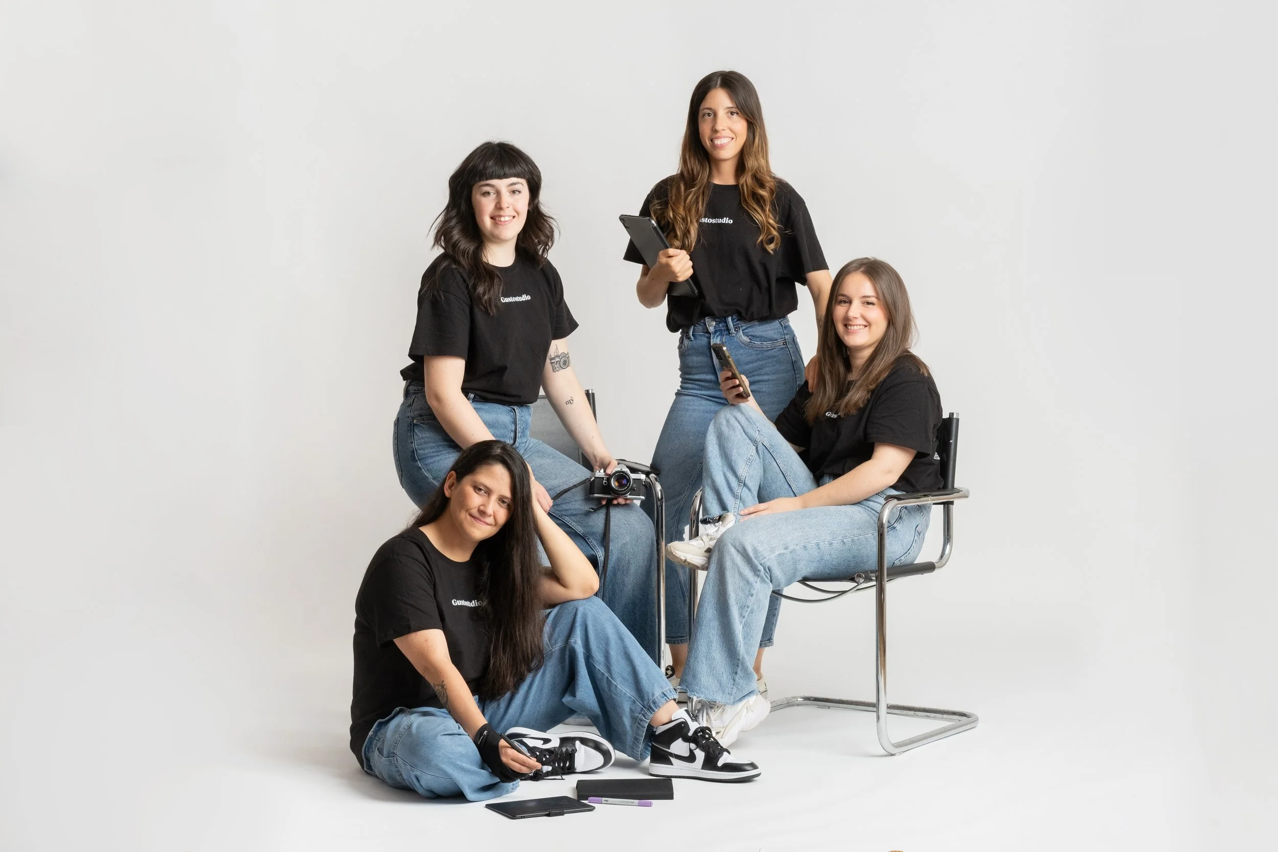 Todo el equipo creativo de GustoStudio, 4 mujeres jóvenes con camisetas negras y jeans en un estudio fotográfico blanco, posando con cámaras y equipo de fotografía.