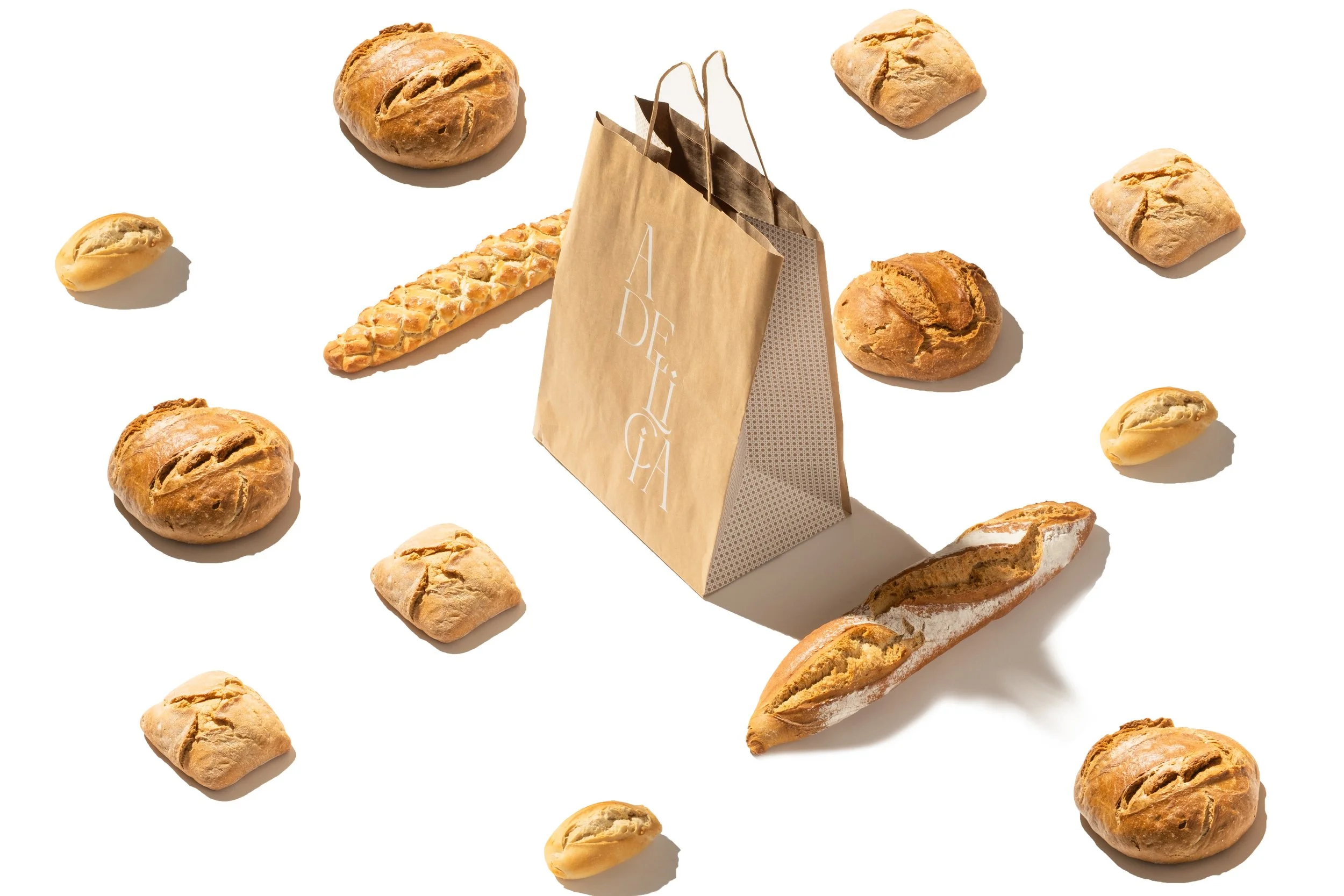 Variedad de panes alrededor de una bolsa de papel que dice 'A DELICIA' en un fondo blanco. Diseño de logotipo y diseño de packaging para una tienda de alimentación en Madrid por GustoStudio. Dirección creativa y foto, también nuestra.