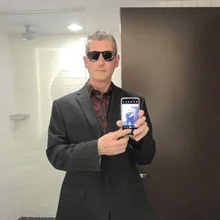 Man taking a selfie in a mirror, wearing sunglasses and a suit in a bathroom.