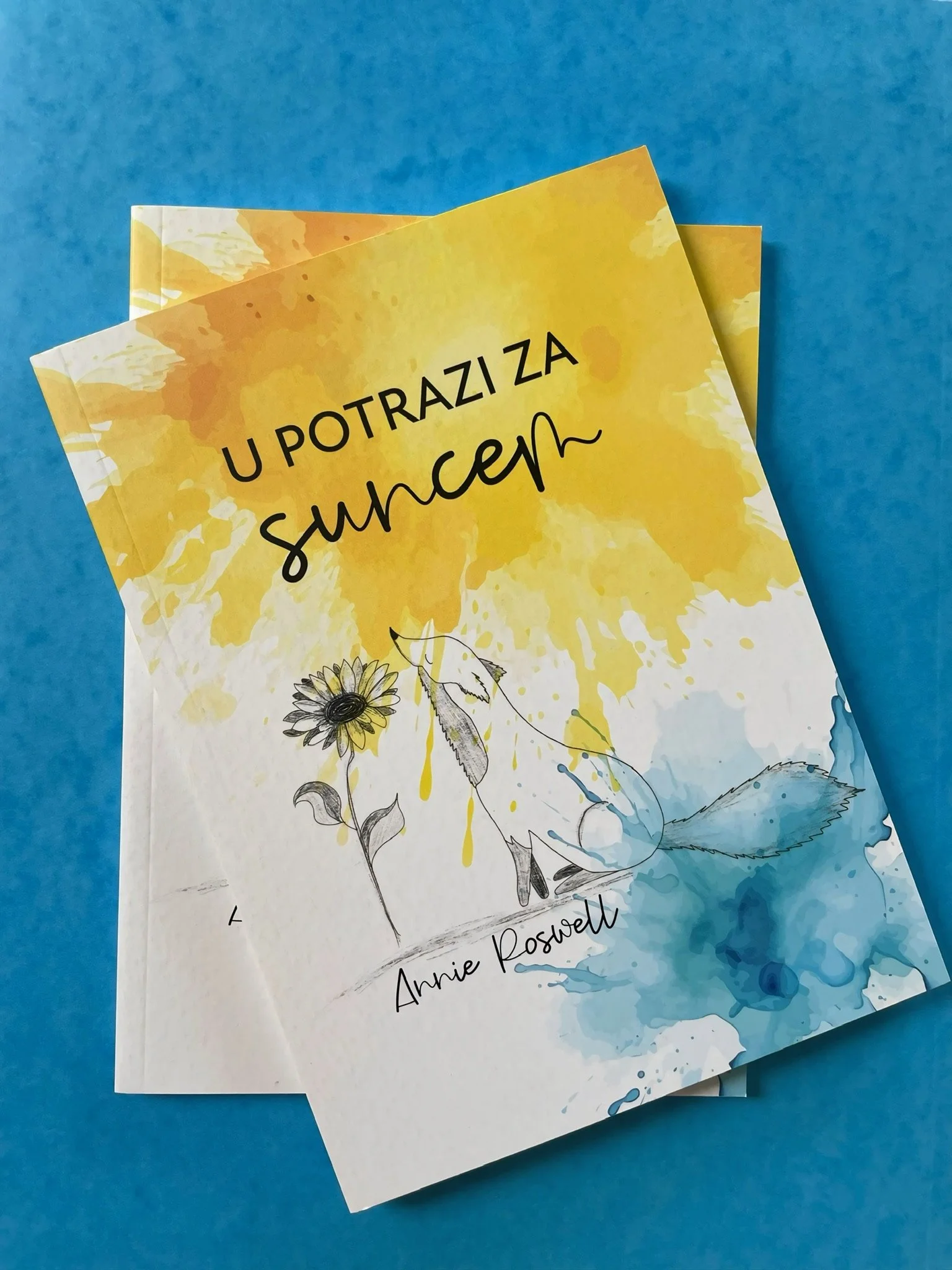 A book titled 'U Potrazi Za Sunceh' by Annie Roswell with a yellow and blue watercolor background, illustrations of a sunflower and a leaf, placed on a blue surface.
