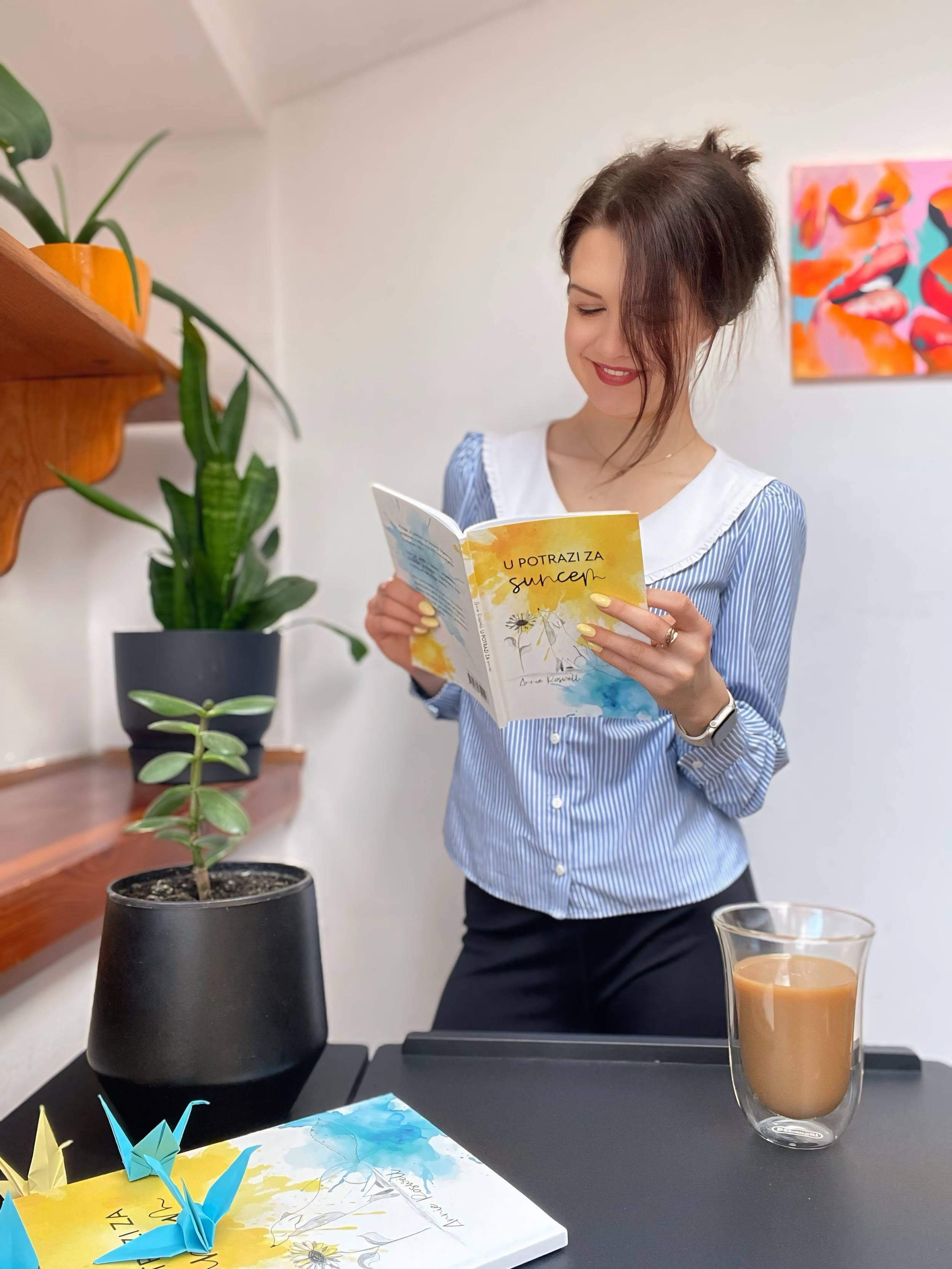 A woman with brown hair in a messy bun, wearing a blue and white striped shirt, is smiling while reading a colorful book titled 'U Potrazi Za Sunce'. She is standing next to a table with a cup of coffee, a potted plant, and origami cranes, in a room decorated with a vibrant abstract painting in the background.