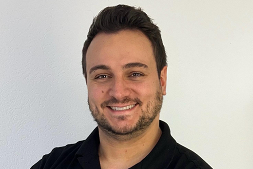 Ein lächelnder Mann mit dunklem Haar und Bart vor einem weißen Hintergrund.