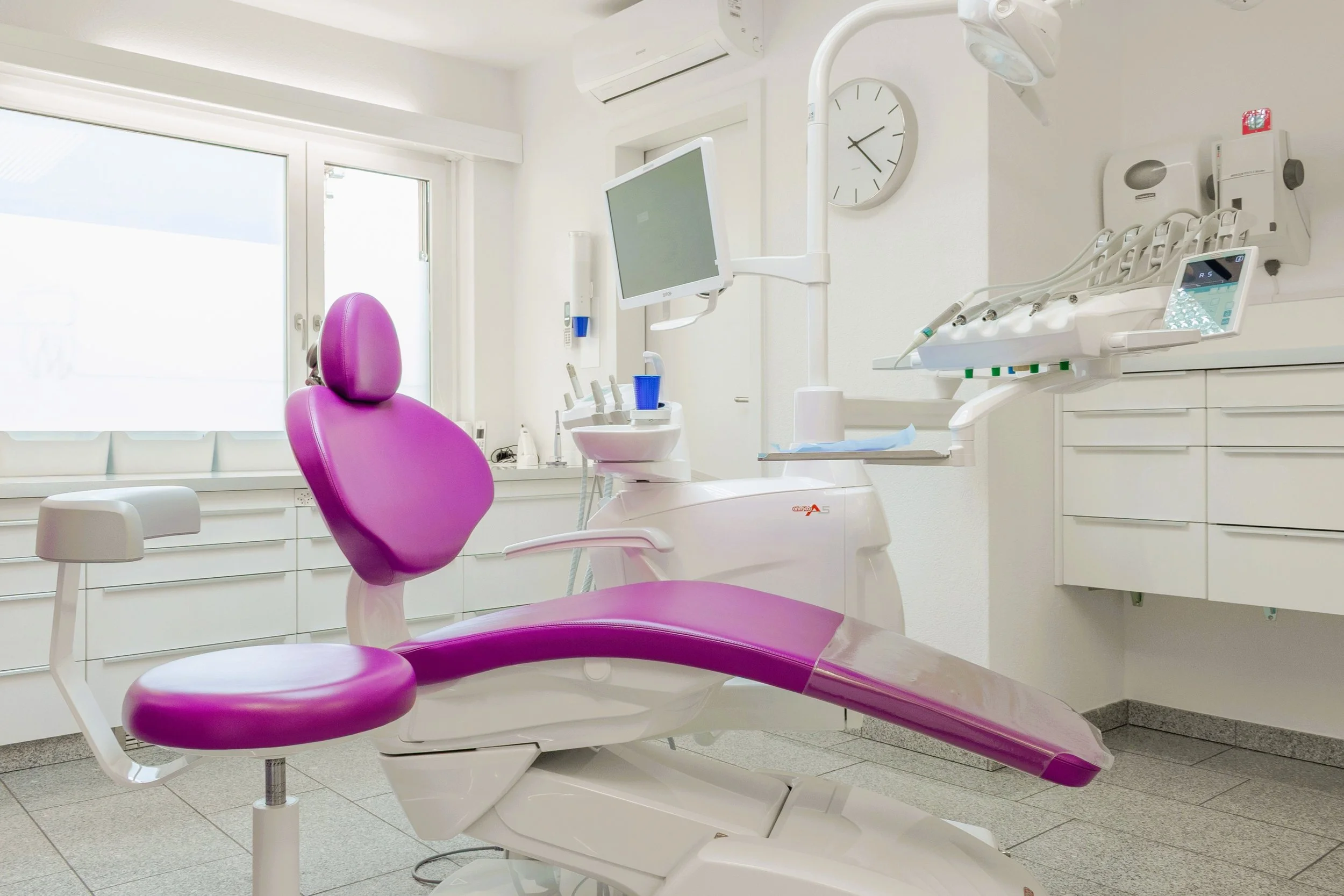 Ein Zahnarztstuhl in einem modernen Behandlungszimmer, umgeben von medizinischer Ausrüstung und Computern.