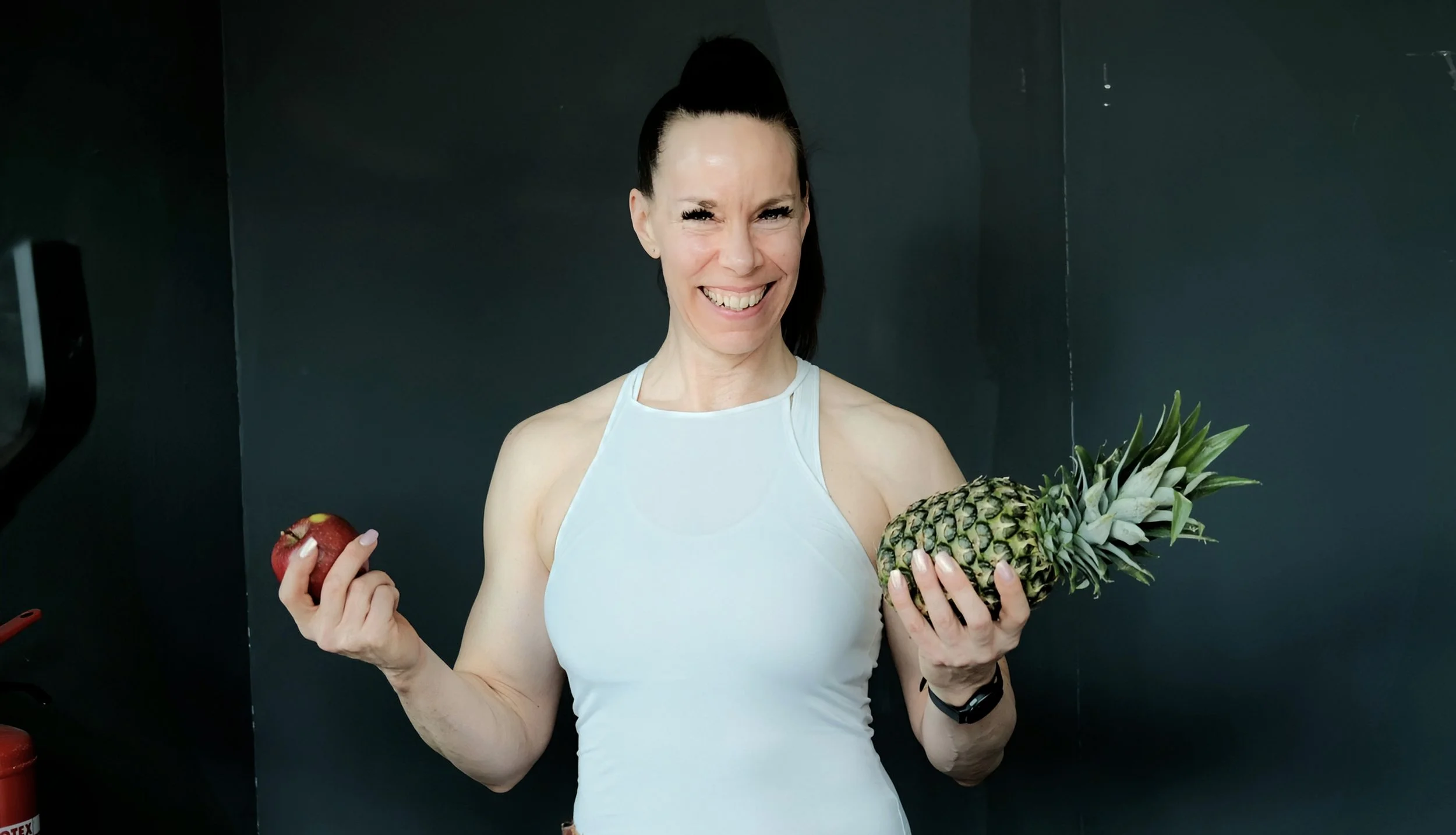 Eine Frau mit schwarzen Haaren in einem weißen Sporttop hält einen Apfel in der linken Hand und eine Ananas in der rechten Hand, sie lächelt in die Kamera vor dunklem Hintergrund.