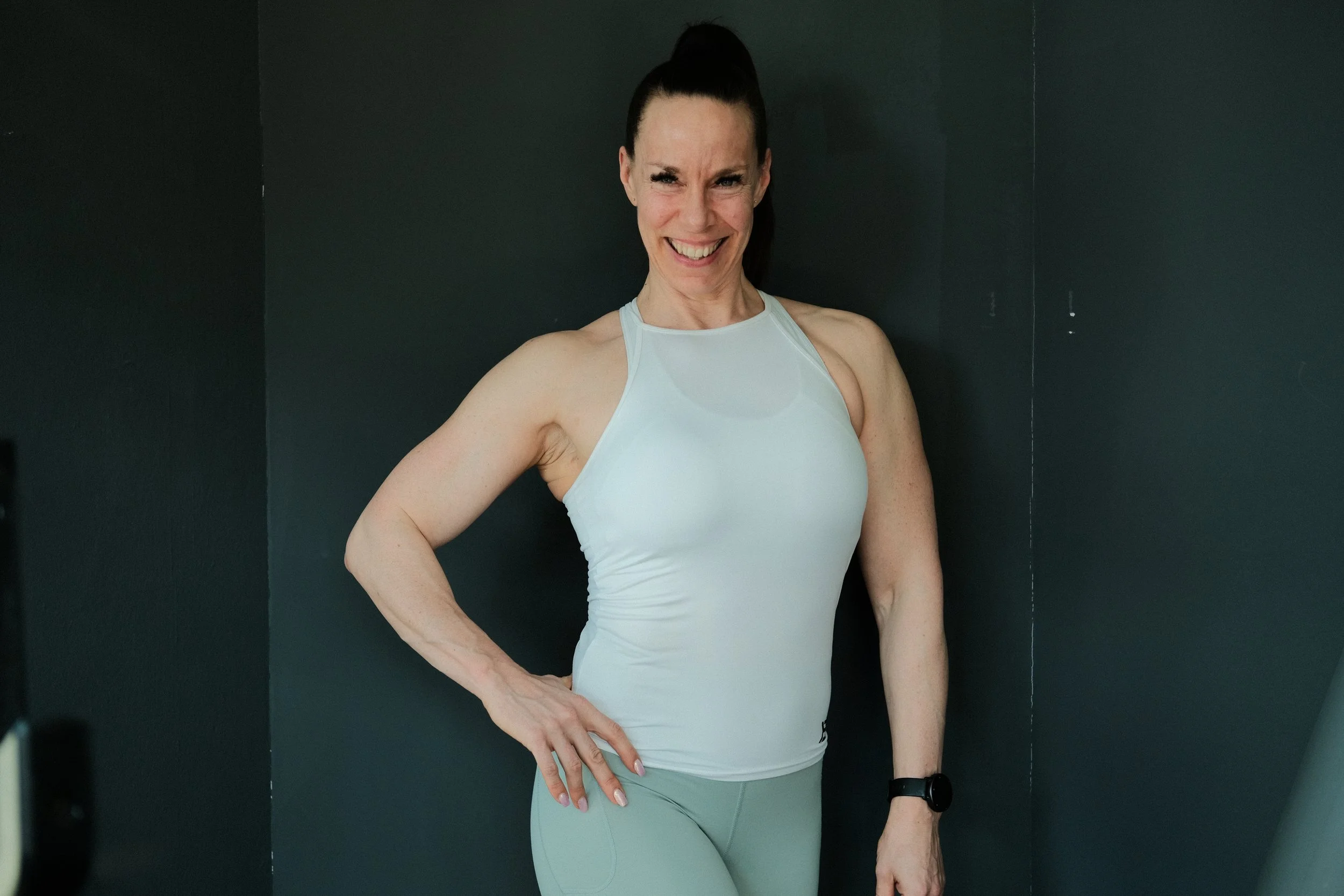 Eine sportliche Frau in weißem Sporttop und grüner Leggings steht vor einer schwarzen Wand, lächelt und posiert mit einer Hand auf der Hüfte.