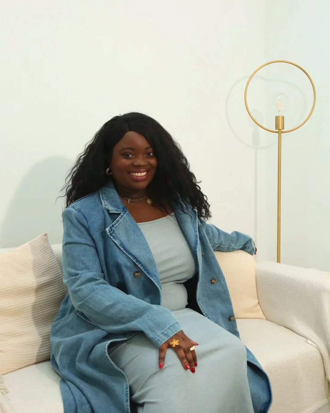 Woman with long hair wearing a denim outfit, sitting on a white couch next to a modern lamp.
