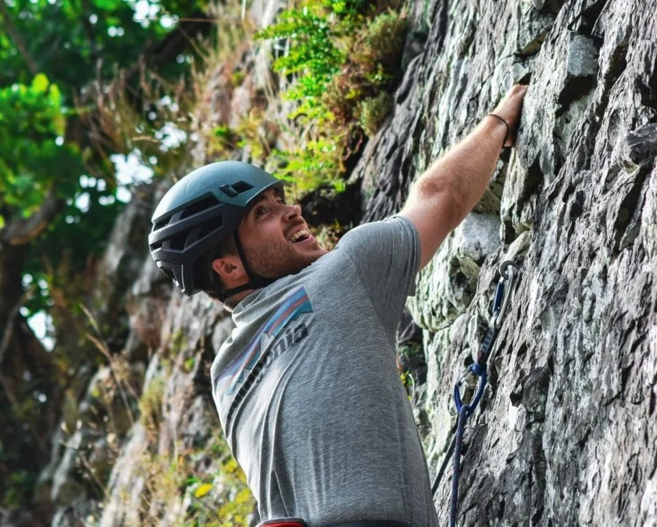 A man climbing a rock wall outdoors, wearing a helmet, reaching upward with his right arm, smiling.