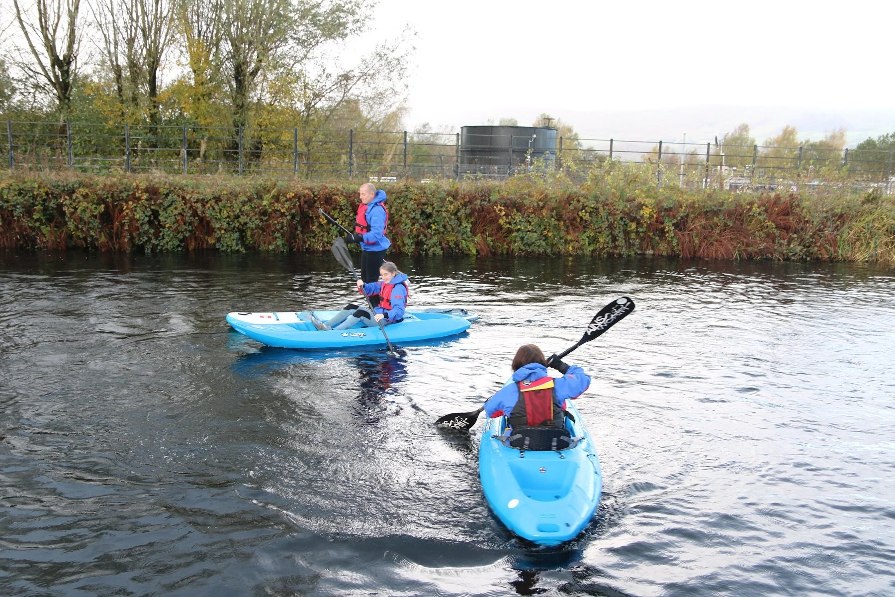 Three people kayaking on a river, two in a blue kayak and one in a blue kayak, with a shoreline of trees and bushes in the background.