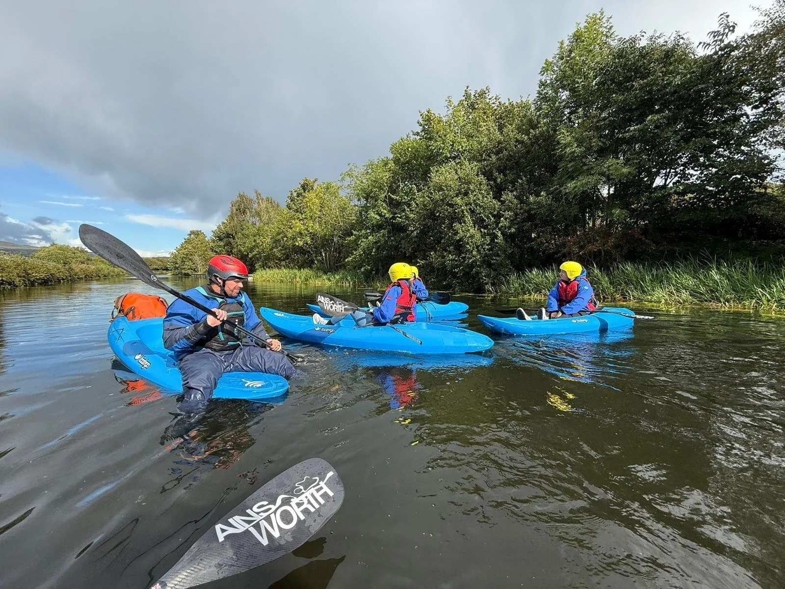 Three people in kayaks on a calm river, surrounded by green trees and bushes, with one person standing in the water holding a kayak paddle.