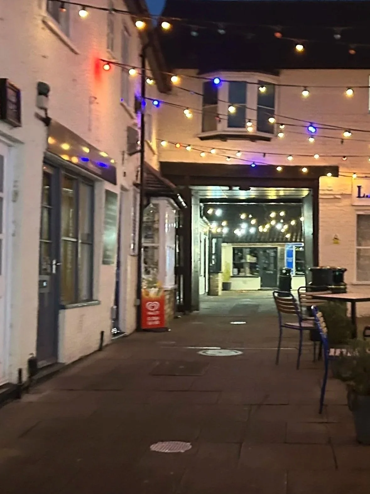 An outdoor patio area decorated with string lights at dusk, featuring tables, chairs, and plants, with a building in the background.