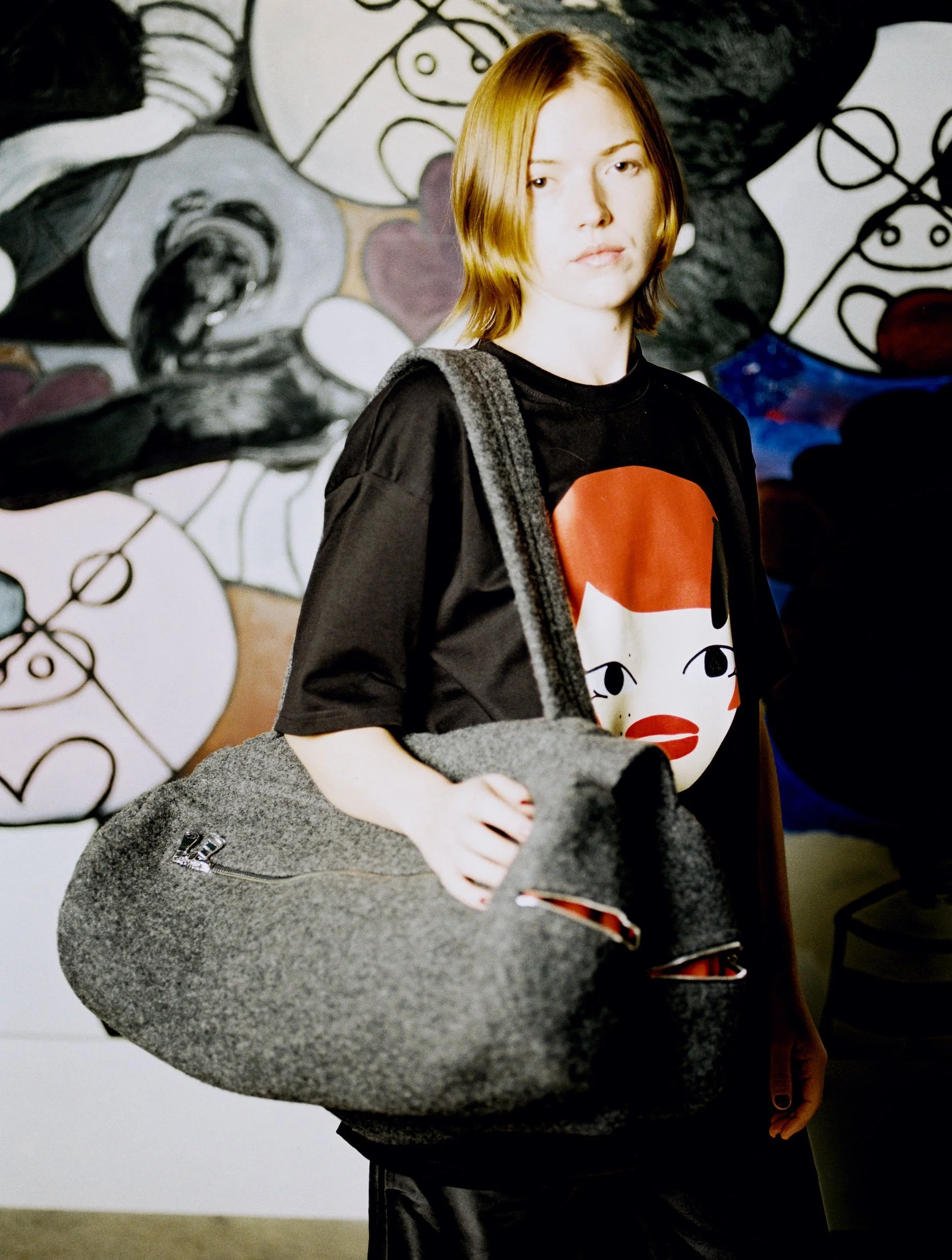 Young woman with shoulder-length red hair, wearing a black t-shirt with a cartoon face graphic, carrying a large gray felt bag, standing in front of a black-and-white abstract mural.