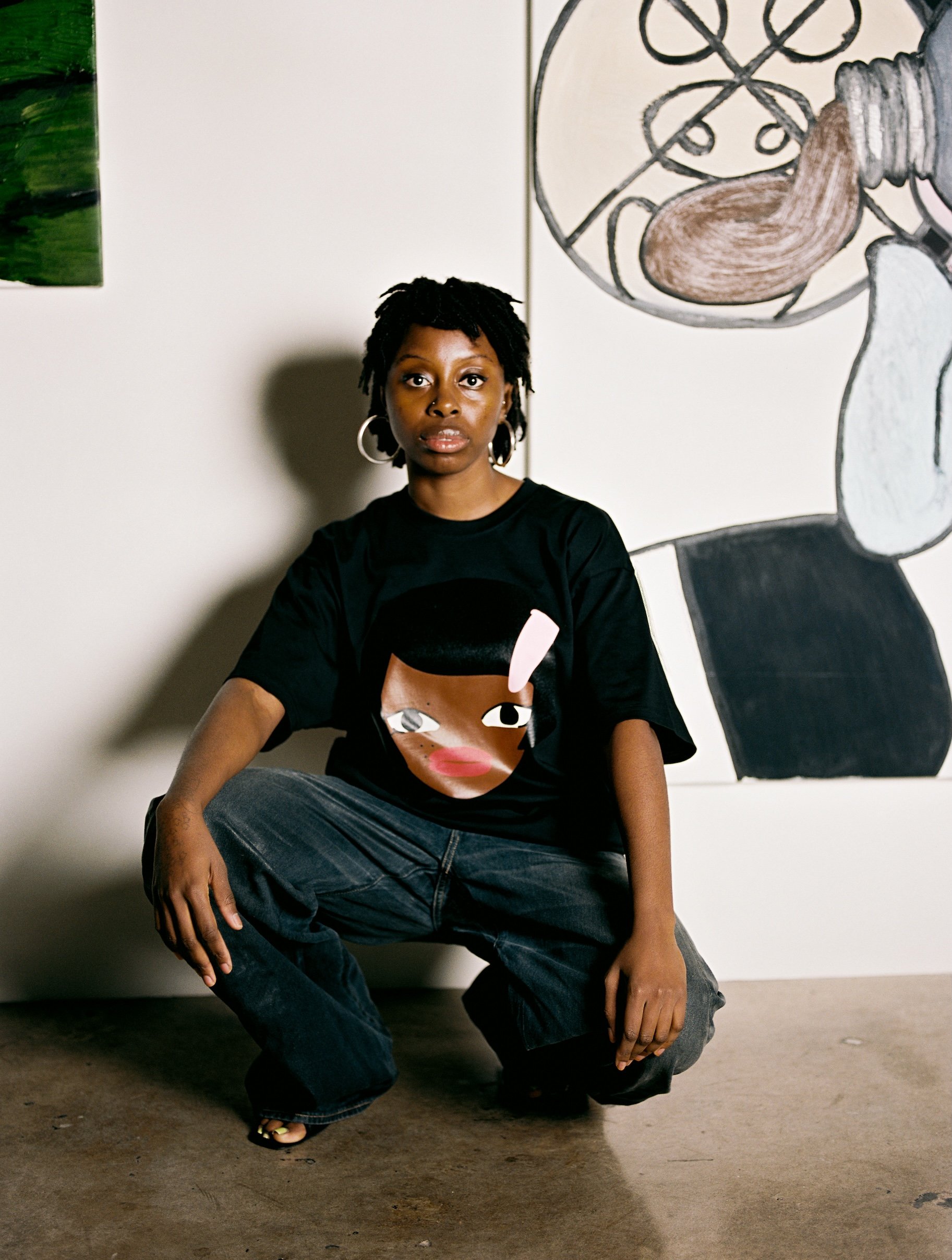 A young woman with short dreadlocks crouching indoors, wearing a black t-shirt with a stylized woman's face on it, in front of abstract artwork on a white wall.