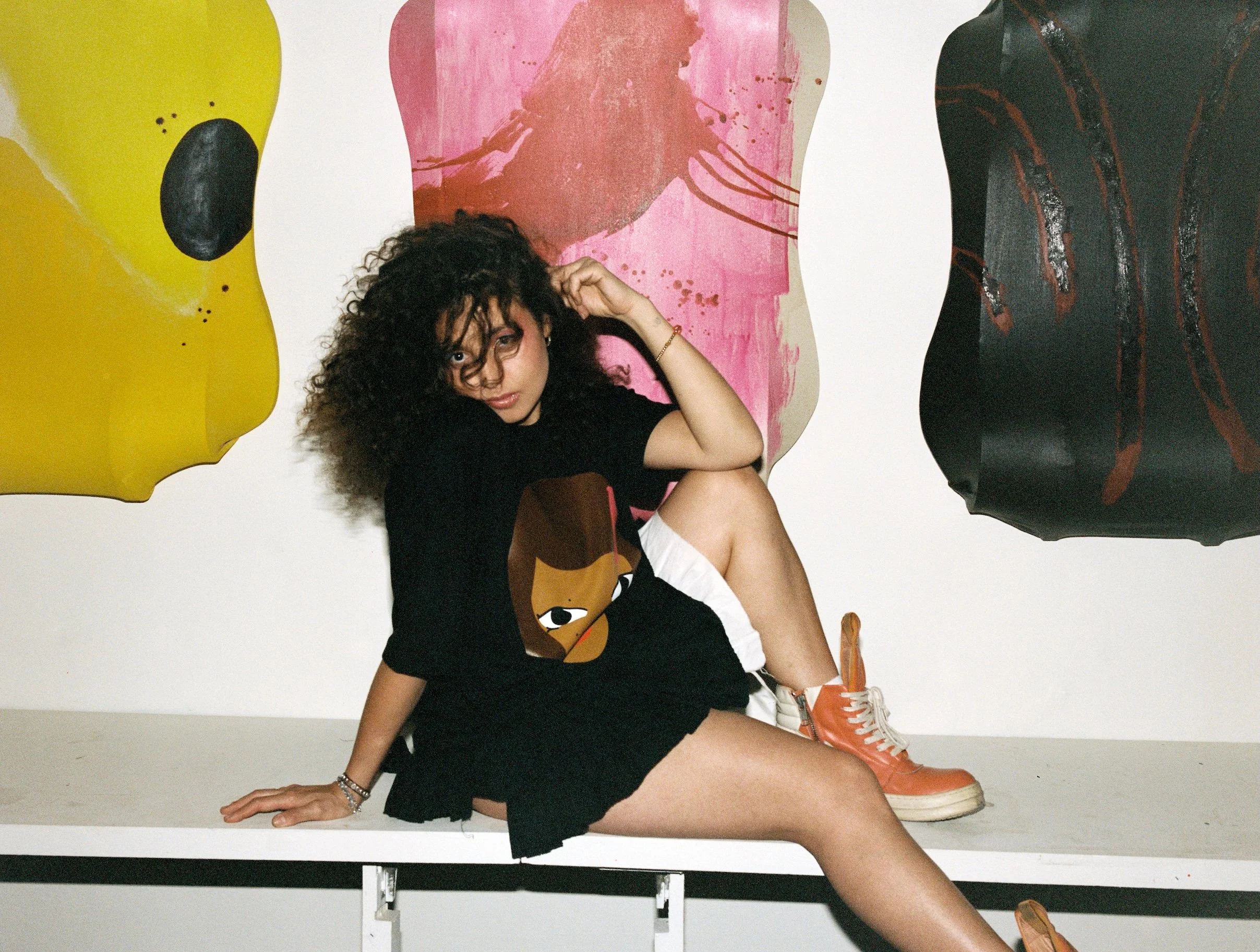 A young woman with curly hair sitting on a white table in front of colorful abstract wall art, wearing a black t-shirt with a cartoon face, white shorts, and orange high-top sneakers.