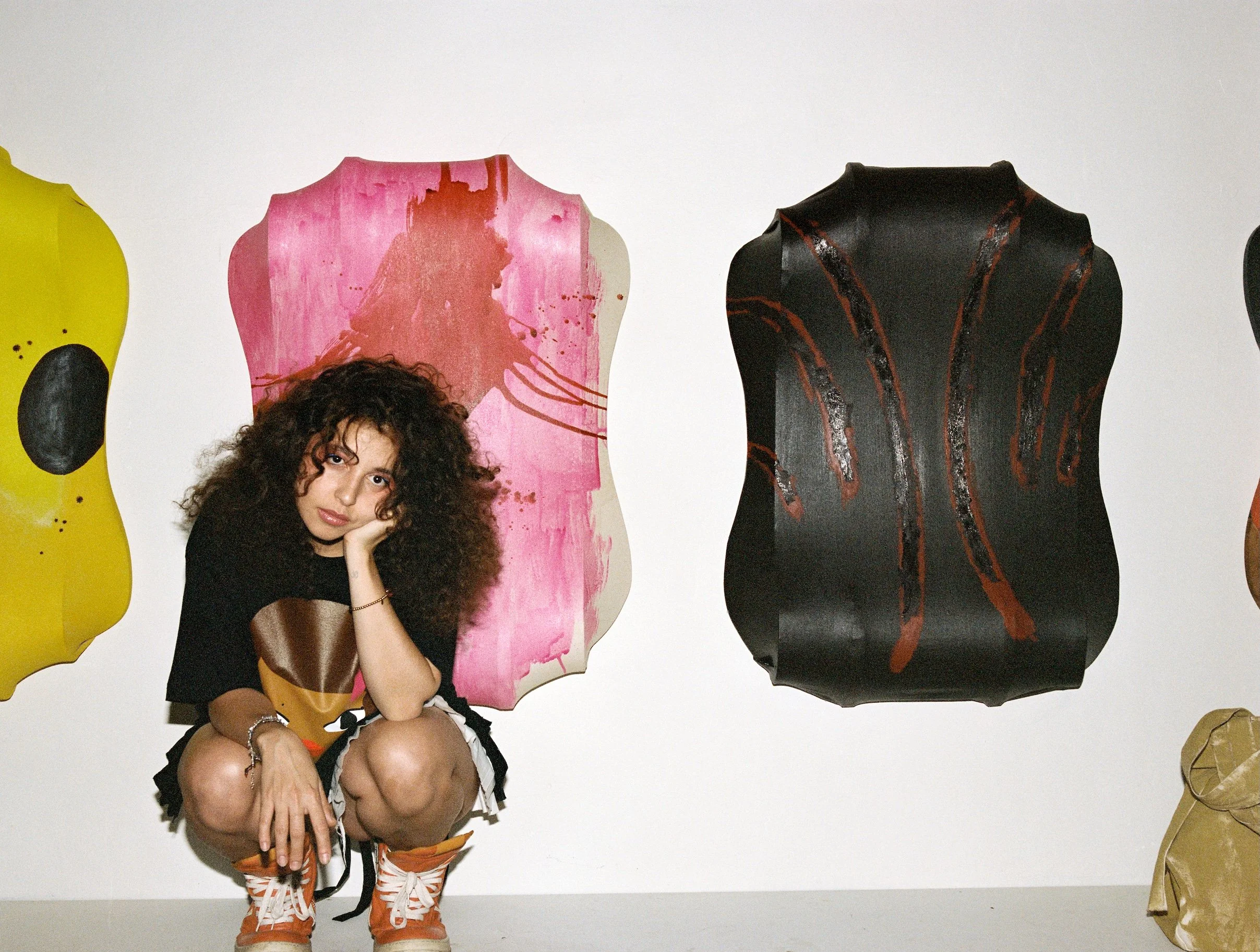 A young woman with curly hair wearing a black t-shirt and colorful shorts squats in front of a plain white wall with abstract, colorful art panels displayed behind her.
